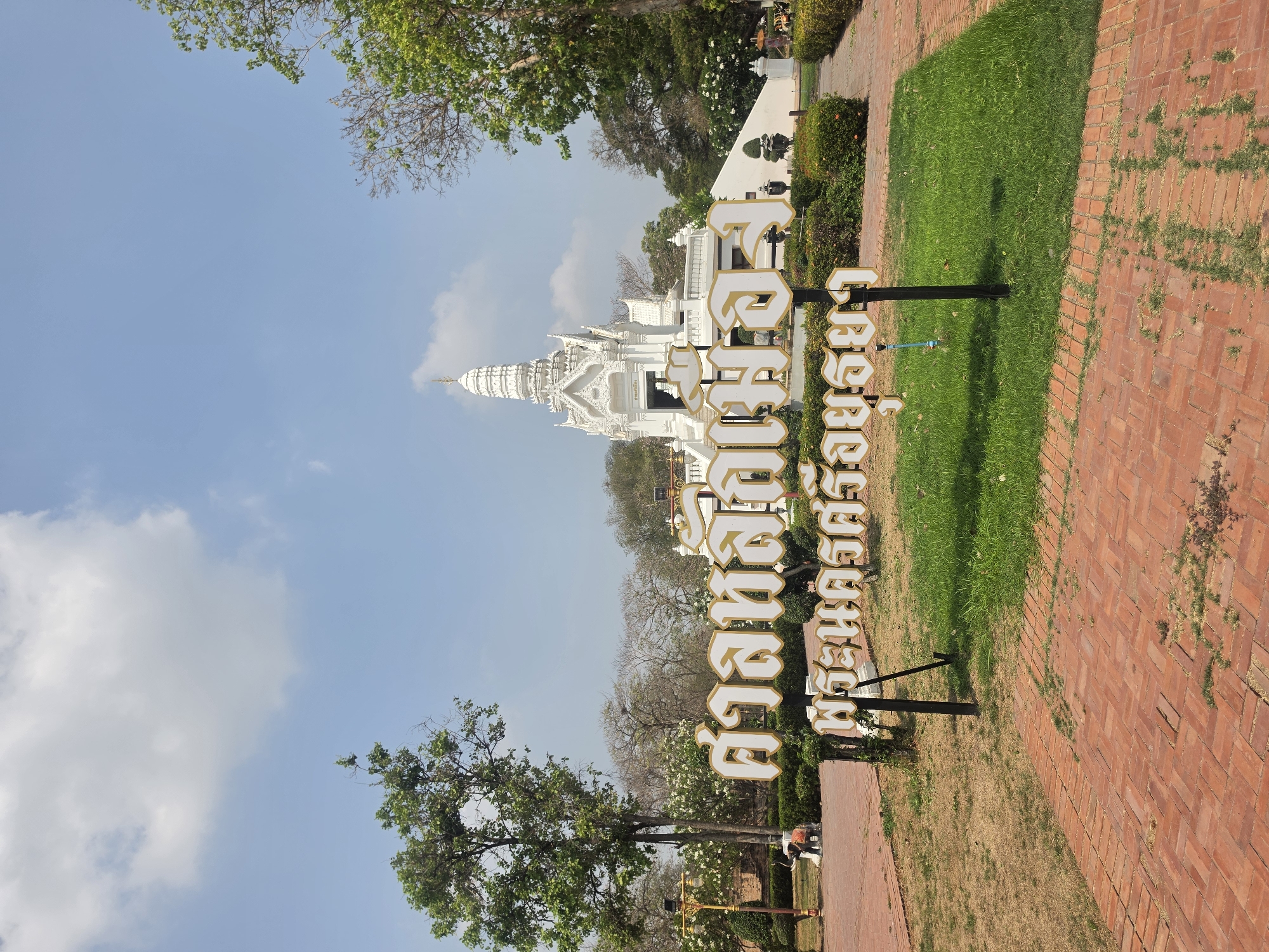 The image shows a white temple building in Historic City of Ayutthaya, Thailand. A sign in Thai characters stands in front of the temple. The surrounding environment consists of trees, grass, and a brick path. The sky is blue with some clouds.
