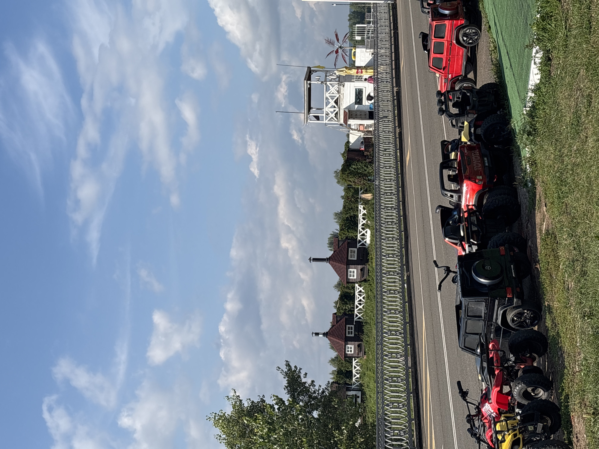 In Naberezhnye Chelny, Russia, a bright, sunny day unfolds with a clear blue sky dotted with white, fluffy clouds. In the foreground, a row of children's ride-on vehicles, including ATVs and miniature SUVs, are parked on a grassy area next to a paved road. Among them, a black toy Mercedes G-Wagon, a red toy Toyota Hilux, and a bright red toy SUV are prominently visible. Beyond the road, a decorative metal fence with an ornate pattern separates the foreground from a backdrop of greenery. Further in the background, several small, rustic-looking wooden houses with red roofs and white-trimmed windows stand amidst trees. To the right, a white observation tower with a lattice design is situated, and a colorful windmill-like sculpture is visible. The overall scene suggests an outdoor recreational area or park designed for children's activities.