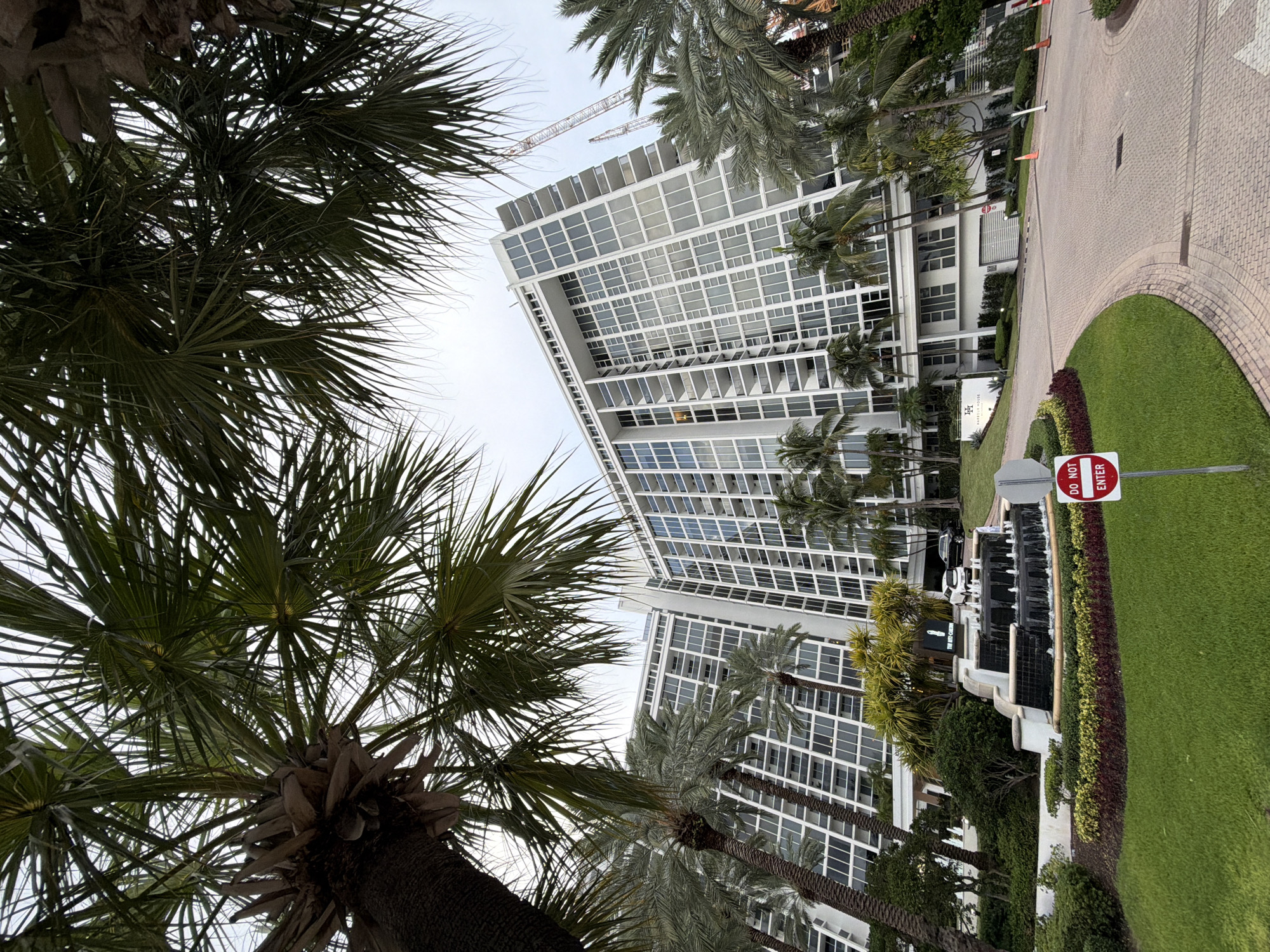 The image captures a frontal view of the Ritz-Carlton Bal Harbour, a luxury hotel located at 10295 Collins Avenue in Bal Harbour Village, Florida, USA. The building is a tall, modern white structure with numerous glass-paneled balconies and windows. In the foreground, palm trees with lush green fronds are prominently featured, some partially obscuring the view of the hotel. A circular driveway leads to the entrance, which is adorned with a multi-tiered fountain and manicured landscaping featuring green grass and colorful bushes. A "Do Not Enter" sign is visible on the right side of the driveway. Two vehicles are parked near the entrance. Construction cranes are visible in the background, suggesting ongoing development in the area. The sky is overcast, and the overall impression is one of a tropical, upscale resort.