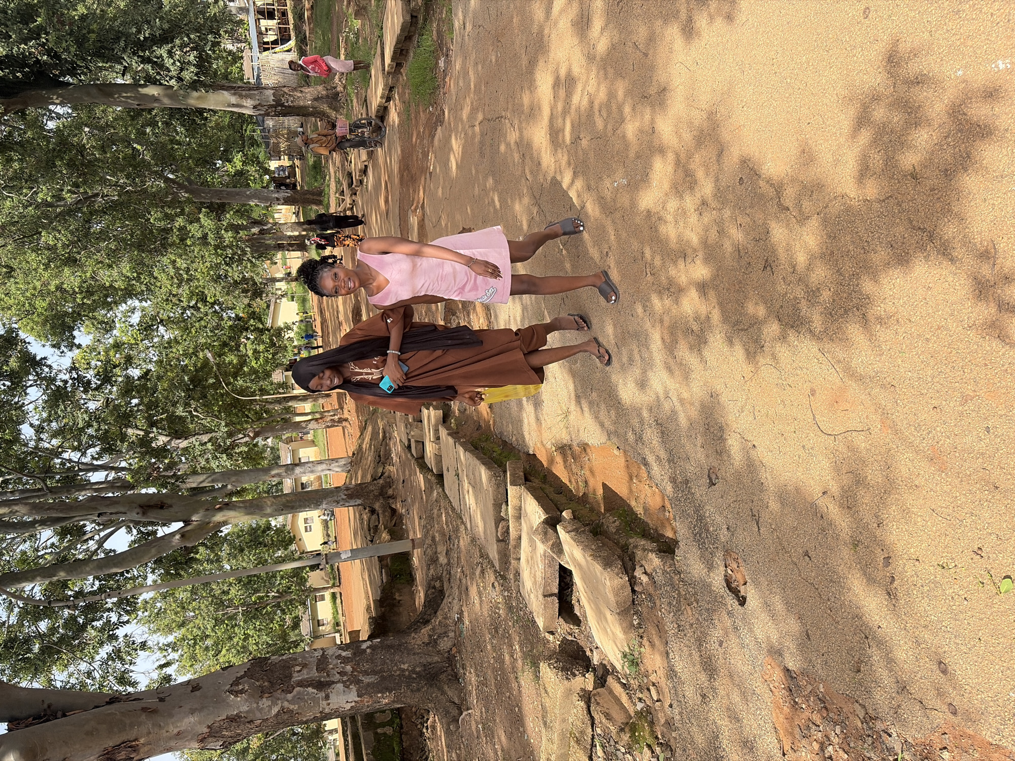 Two young women are standing on a dirt path. The woman on the left is wearing a brown, ankle-length dress and a black headscarf. She is holding a blue smartphone in her right hand. The woman on the right is wearing a short, light pink dress and flip-flops. Both women are smiling. They are likely outside, given the presence of trees and sunlight casting shadows on the path. The setting appears to be a somewhat rural or semi-urban area with buildings visible in the background and a line of concrete slabs running alongside the path. The overall atmosphere seems relaxed and pleasant. The time of day appears to be daytime, with bright sunlight. The location is Rafin Par, Nigeria.