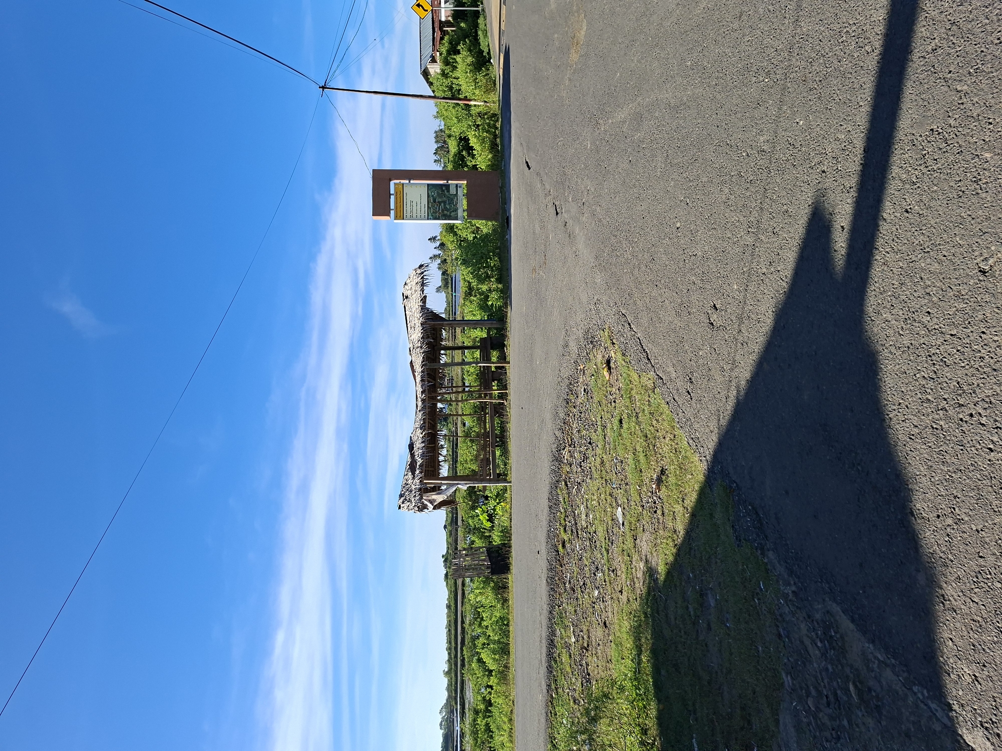 The image shows a roadside scene in Kutaglumpang, Indonesia. A small, thatched-roof structure stands next to a two-lane paved road. A sign with informational text and images is posted nearby. Beyond the structure, there is green vegetation and a blue sky with wispy clouds. A shadow is cast on the road's surface. Power lines are visible against the sky.