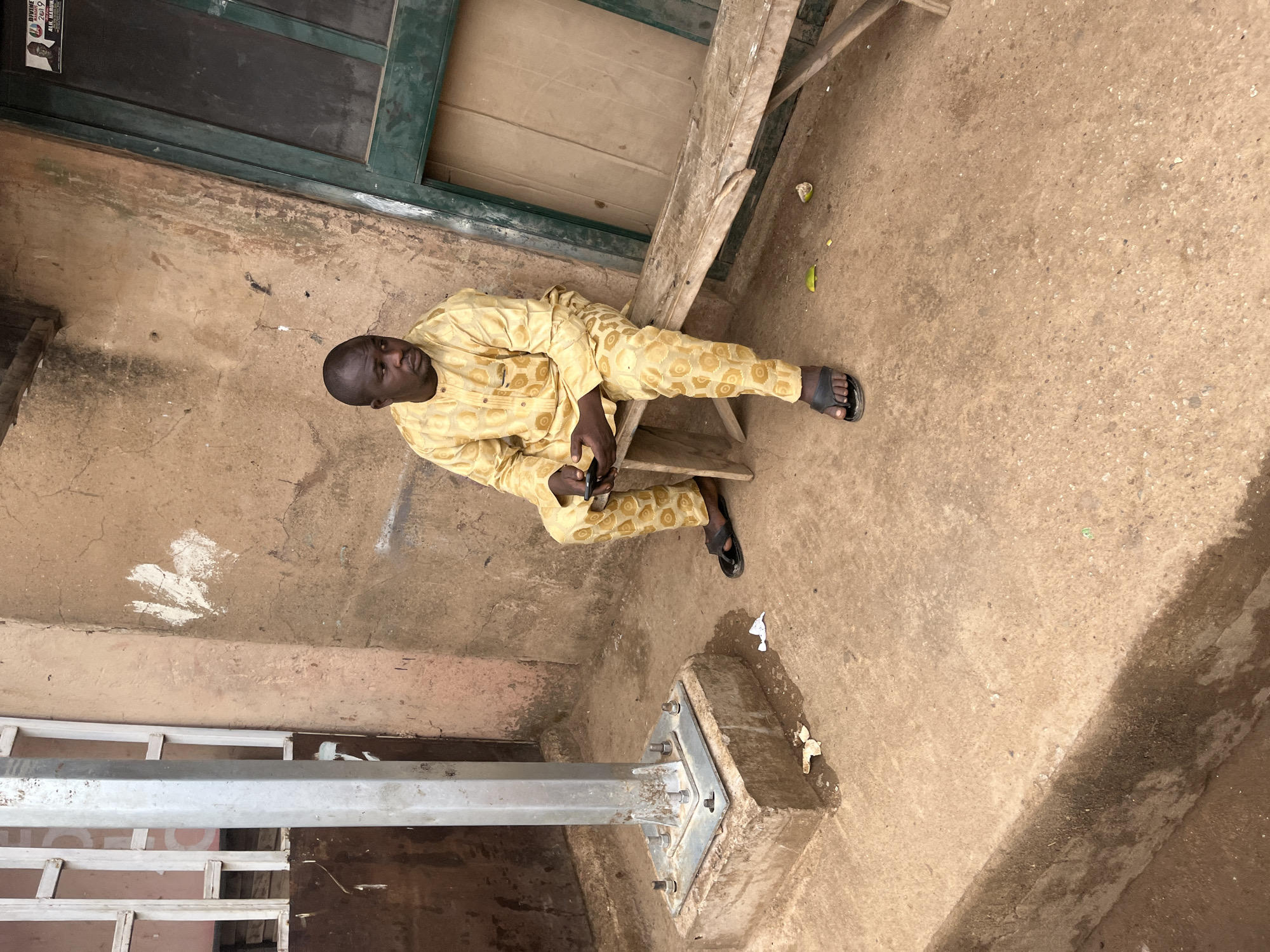 A man in a patterned yellow outfit and black sandals sits on a wooden bench outside a building in Jos, Nigeria. The building has a green-framed window. The man holds a dark-colored object, possibly a phone, in his hands. A gray metal pole stands in the foreground, anchored to a concrete base. The ground is dirt.