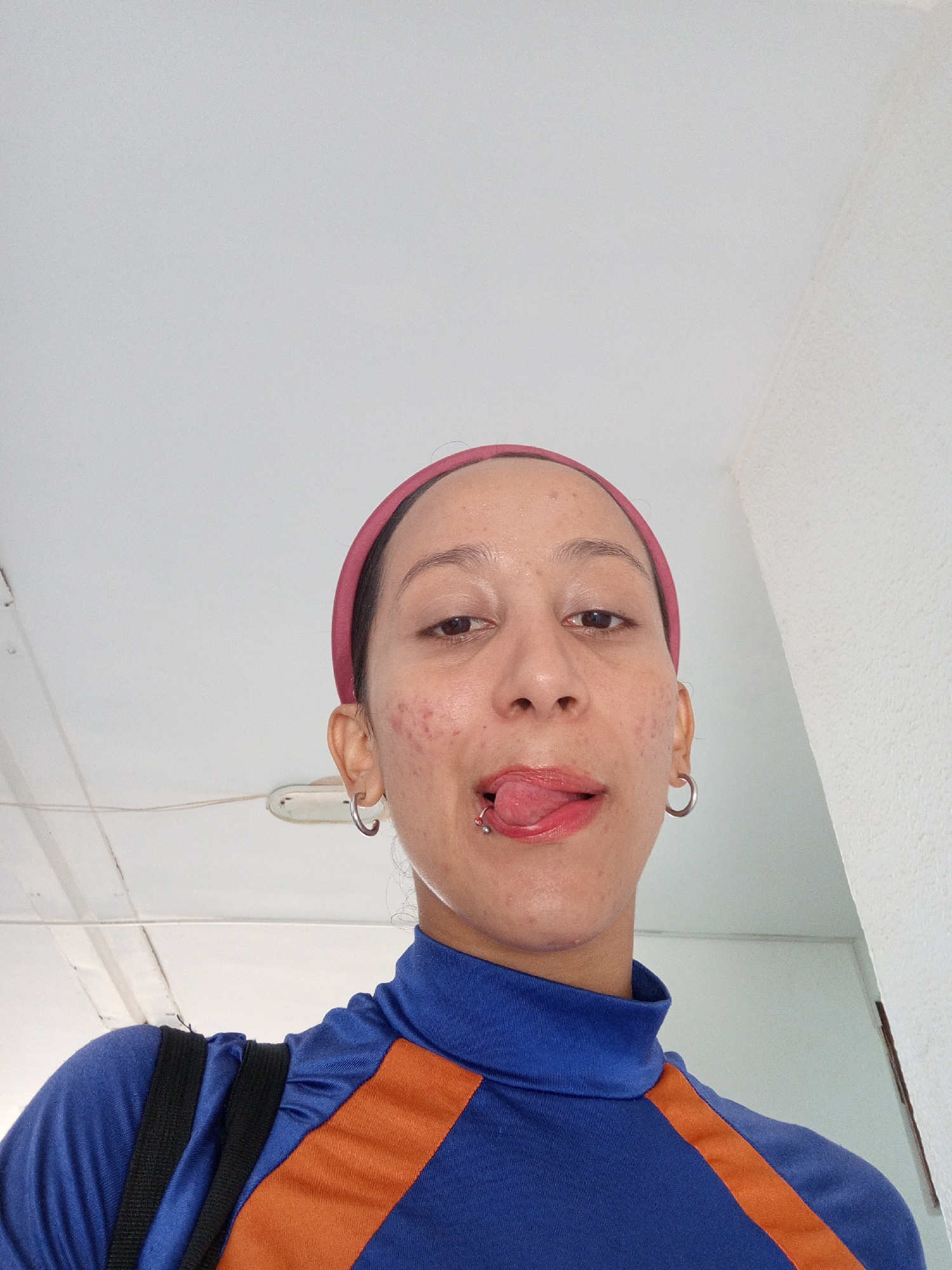 A selfie was taken by a person with dark hair and a pink headband, wearing a blue and orange turtleneck shirt with black straps over their shoulder. They have pierced ears with hoop earrings, a pierced tongue with a barbell, and are sticking their tongue out. The background includes a white ceiling, a fluorescent light fixture, and a white wall. The location is identified as Porlamar, Venezuela.