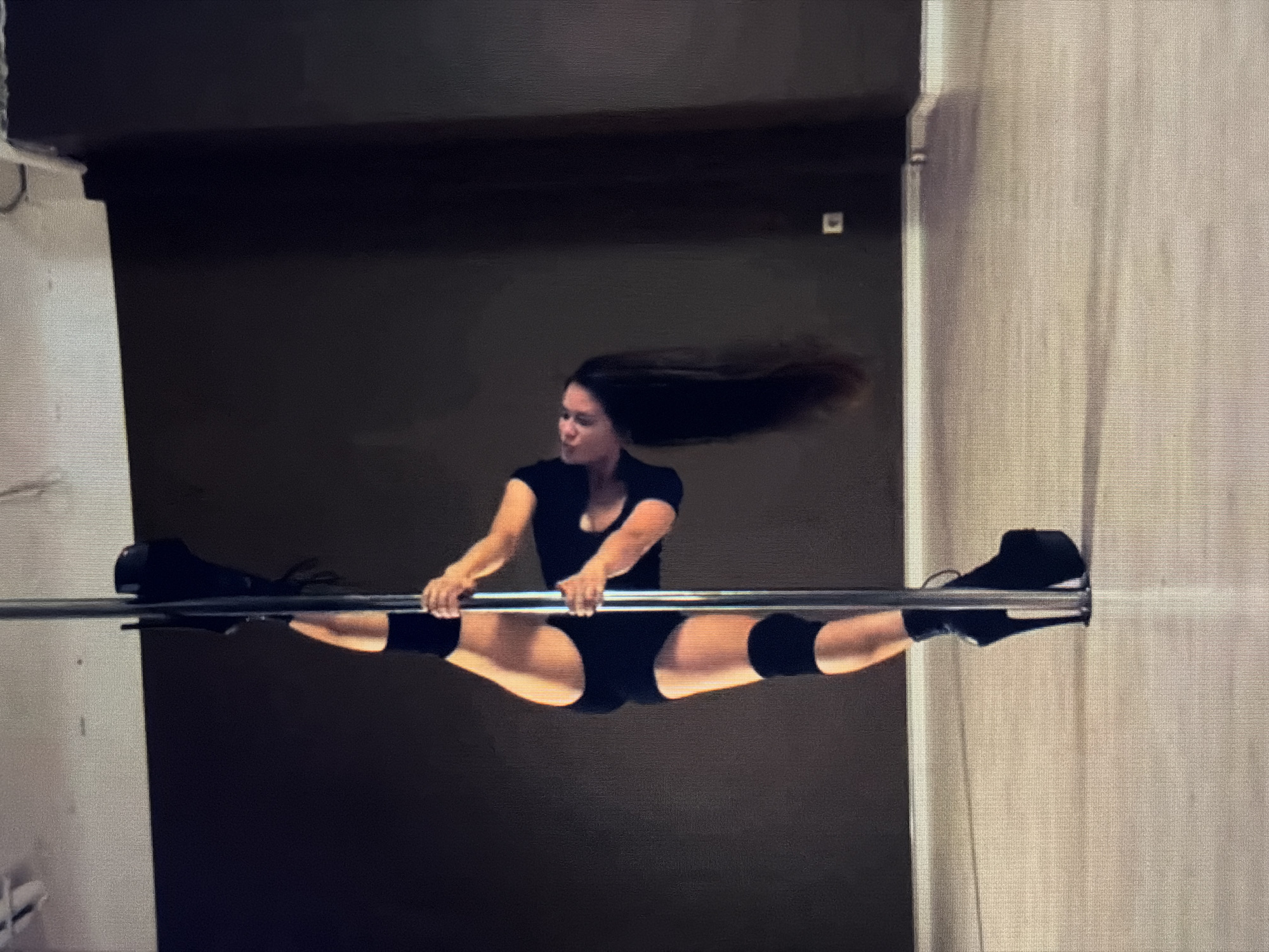 A woman is performing a pole dance in a studio. She is wearing a black leotard, black knee pads, and black platform boots. Her body is positioned in a vertical split, with one leg extended upwards and the other bent at the knee, wrapped around the pole. She is holding onto the pole with both hands and her head is tilted back. The background is a dark brown wall and a light-colored floor. There is a white ceiling with some lighting fixtures visible. The image appears to be taken from a slightly low angle. There are no visible signs or indications of the location being in the city of Sirius, Russia.
