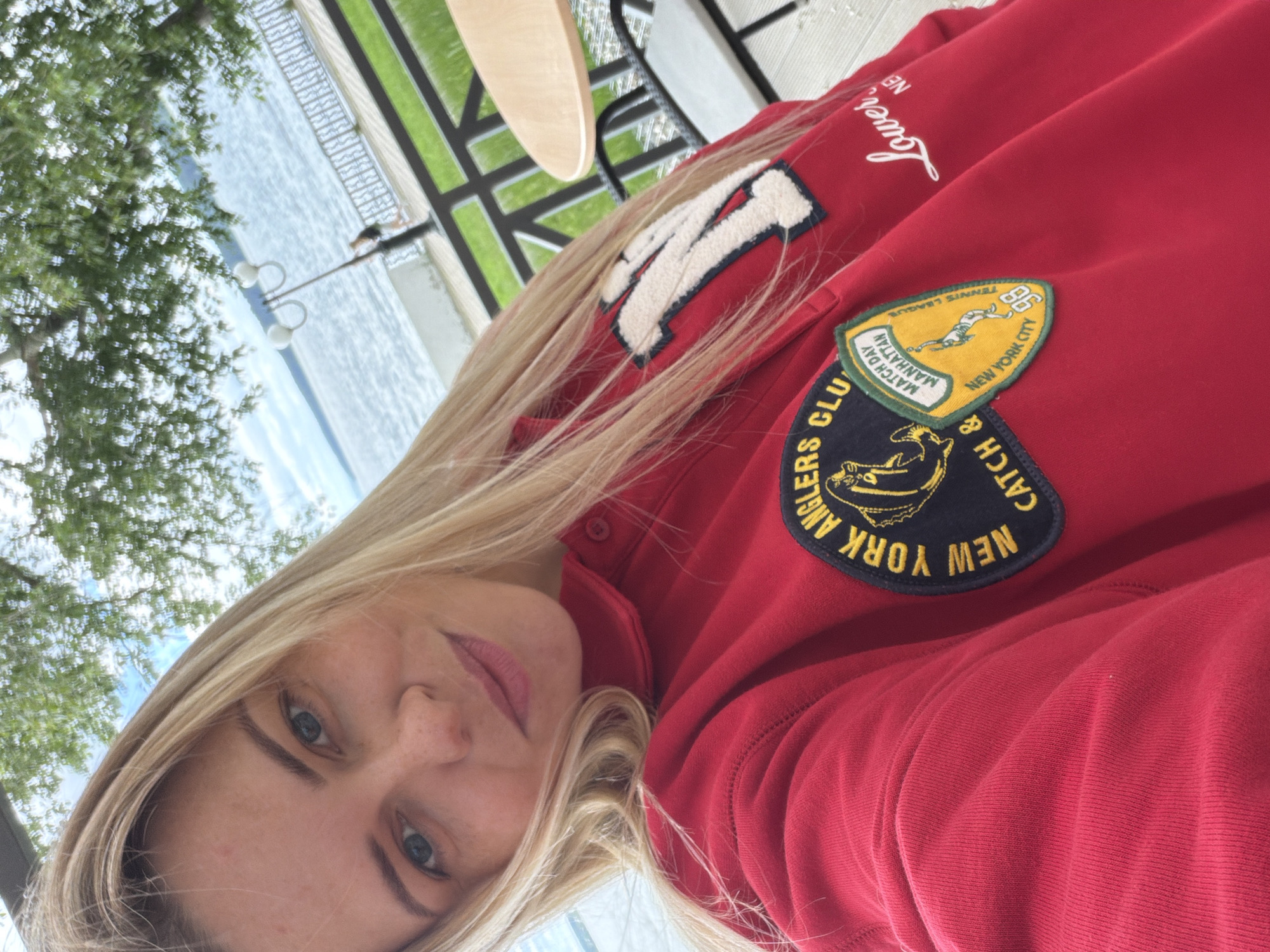 A young woman with blonde hair and blue eyes takes a selfie outdoors. She is wearing a red sweatshirt with patches on it. One patch says "NEW YORK ANGLERS CLUB CATCH &". Another patch features a yellow shield with a graphic of a tennis player and text that reads "MATCH DAY MANHATTAN NEW YORK CITY TENNIS LEAGUE 98". A third patch is on the upper left side of the sweatshirt, with the letter "M" in white outlined in blue, surrounded by a cream-colored fuzzy border. She is sitting at a table near a body of water, possibly a lake or river. In the background, there is a railing, a lamppost, and trees. The sky is visible with clouds. The woman is looking at the camera and smiling slightly.