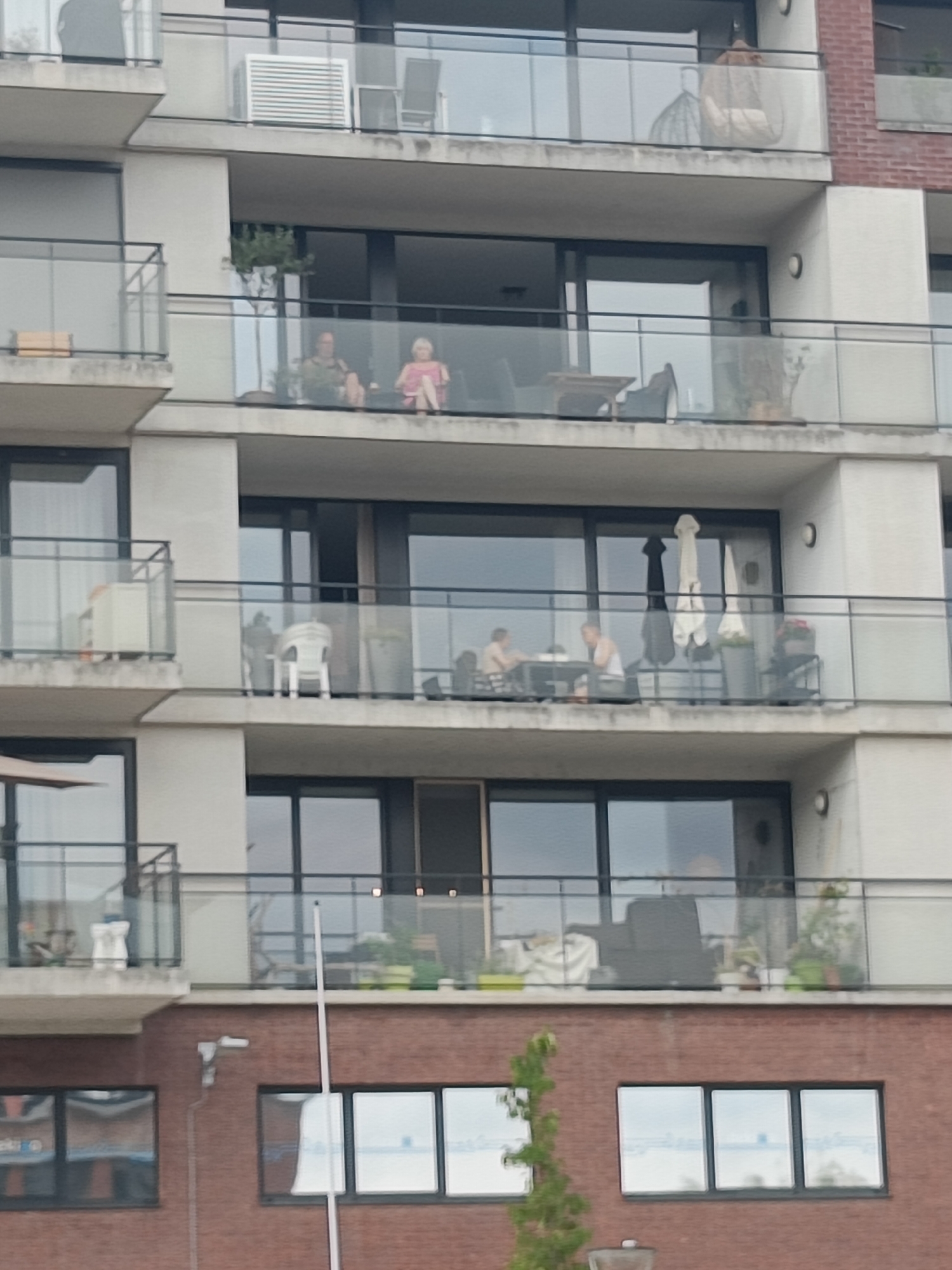 The image shows a multi-story apartment building with balconies. On the third-floor balcony from the top, two individuals are visible sitting on chairs. One is a person wearing a pink top and light-colored bottoms, and the other is a person wearing a darker top and bottoms. There is a small tree on this balcony. The building is constructed of concrete and brick. The setting appears to be urban.