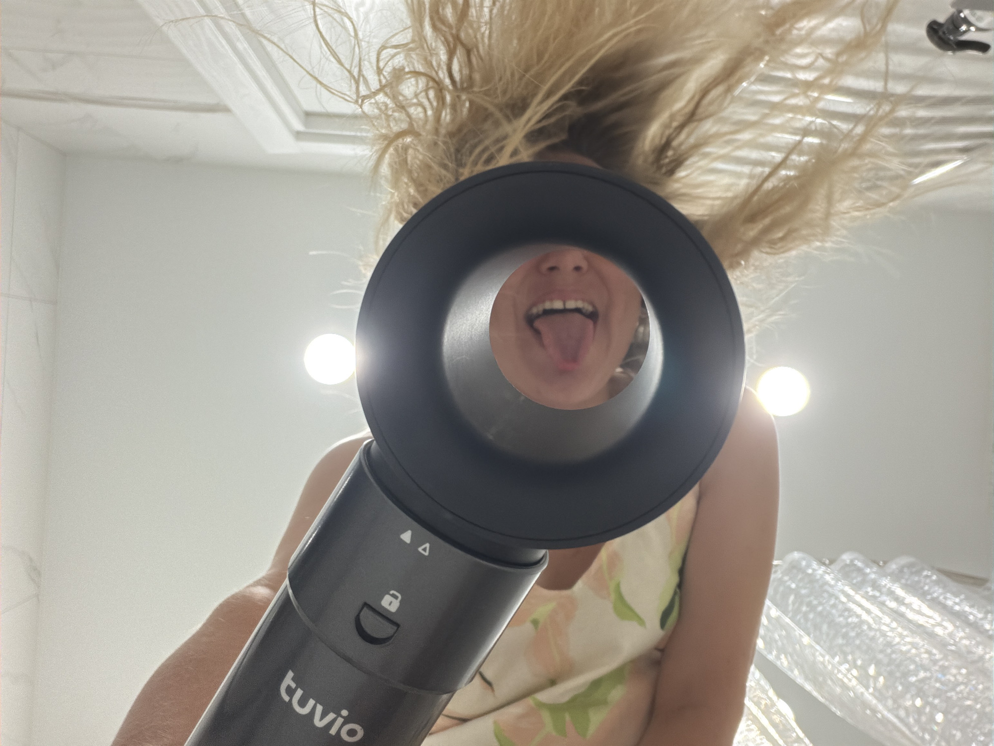 A person, seen from a low angle looking up, holds a dark gray hairdryer with "tuvio" printed on it. The hairdryer is pointed towards the viewer, and through its opening, a person's open mouth with their tongue sticking out is visible. The person holding the hairdryer has blonde hair and is wearing a floral patterned top. The environment appears to be a bathroom, with white tiled walls and bright lighting. There is a shower curtain or similar translucent material visible at the bottom of the image. The overall scene is a close-up, distorted perspective.