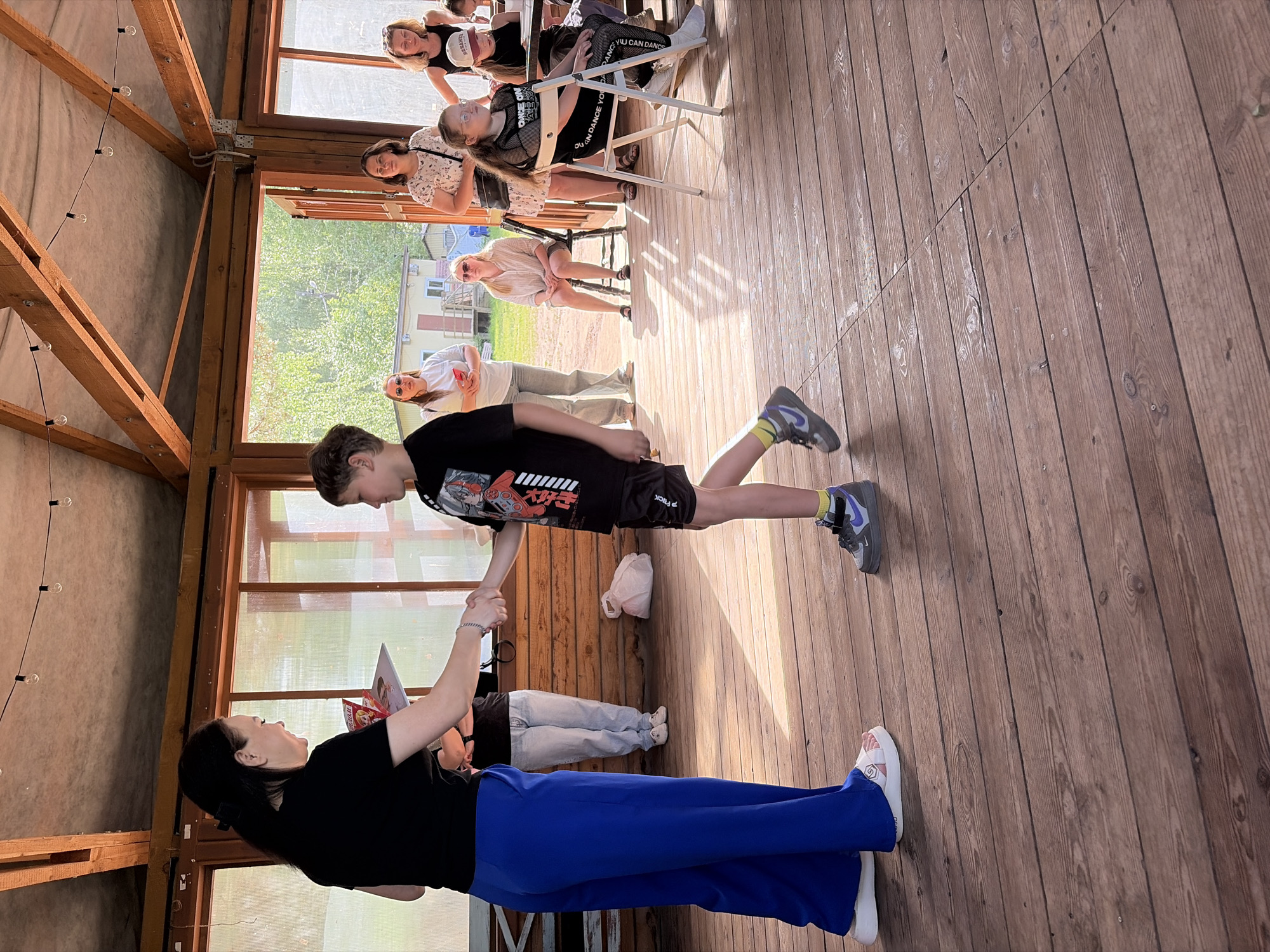 A group of people are in what appears to be a warm wooden cabin structure with open door spaces that lead to an outdoor area. People can be seen sitting on chairs in a row, with others standing and interacting. One woman in a black top and bright blue pants is holding a booklet while shaking hands with a younger boy wearing a graphic tee and sneakers. The location may be in Chaikovsky, Russia.