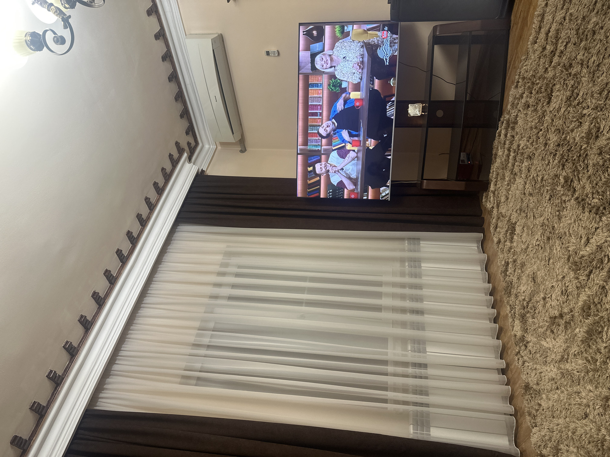 The image shows an interior scene, possibly a living room, with a television displaying a show with three people. The TV is mounted on a stand, and there is a rug on the floor. The room has brown and white curtains, and an air conditioning unit is visible on the wall. Based on other information, the location is Yerevan, Armenia.