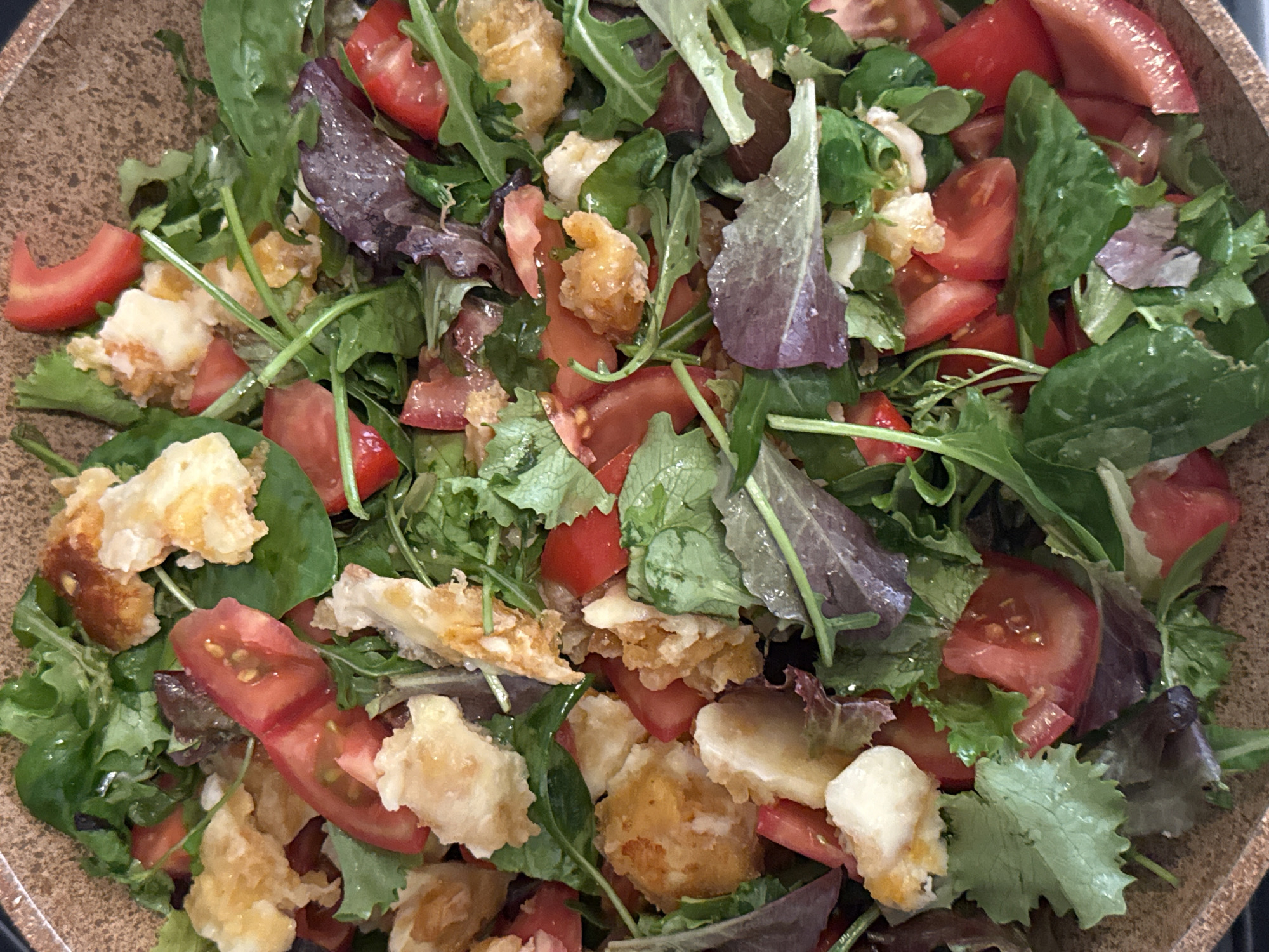 The image shows a close-up view of a salad in a brown bowl. The salad consists primarily of mixed greens, sliced tomatoes, and what appears to be fried cheese or croutons. The greens include various types of lettuce, some with a reddish hue. The tomatoes are cut into wedges. The fried cheese/croutons are irregularly shaped and golden brown. There is no indication of the location of Bar, Montenegro.