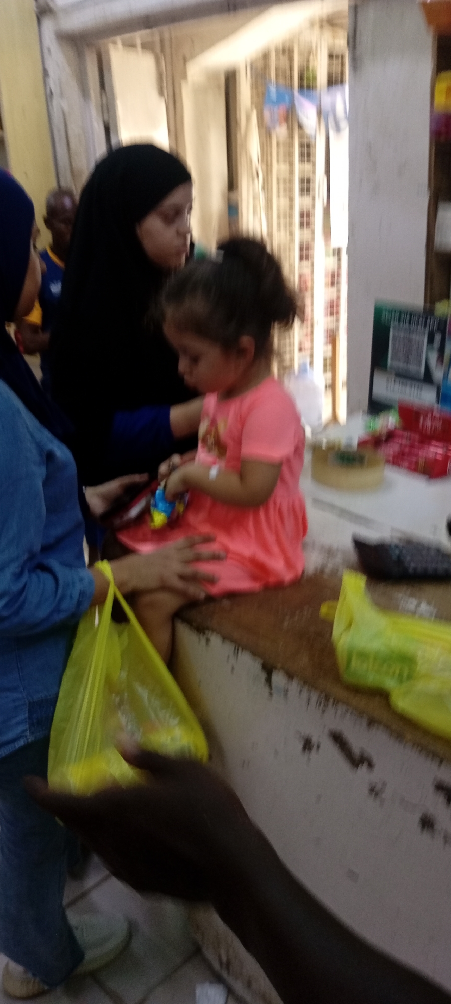 A medium shot captures a scene in Tudun Wada, Nigeria. A young girl with her hair in a bun sits on a ledge, dressed in a peach-colored dress and playing with a blue and yellow toy. To her left stands a person in a blue denim shirt, holding a yellow plastic bag. Behind the child, a woman in a black hijab looks down at her. In the background, another person with dark skin is visible. The ledge upon which the girl sits appears to be a counter of some sort, with various items scattered on top. A hand with dark skin reaches into the frame from the bottom. The setting appears to be inside a building, possibly a shop, indicated by the presence of what looks like retail items.