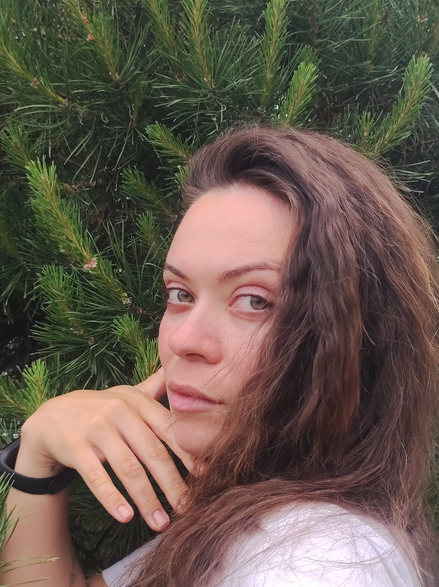 A medium close-up shot captures an adult female from the chest up, positioned against a dense background of dark green pine foliage. The woman has long, wavy, dark brown hair that falls over her shoulders and partially obscures the right side of her face. Her skin tone is fair, and her eyes, appearing green or hazel, are directed straight at the viewer with a serious expression. Her nose shows a slight redness on the bridge/tip. Her right hand is raised, with fingers gently touching her chin/jawline on the left side of her face. She wears a white t-shirt, and a dark, possibly black, fitness tracker or watch is visible on her right wrist. A faint dark mark or tattoo is discernible on the inner part of her right forearm. The pine needles in the background are sharp and vibrant green, some with lighter tips indicating new growth, forming a uniform, natural backdrop in the city of Unknown, Unknown.