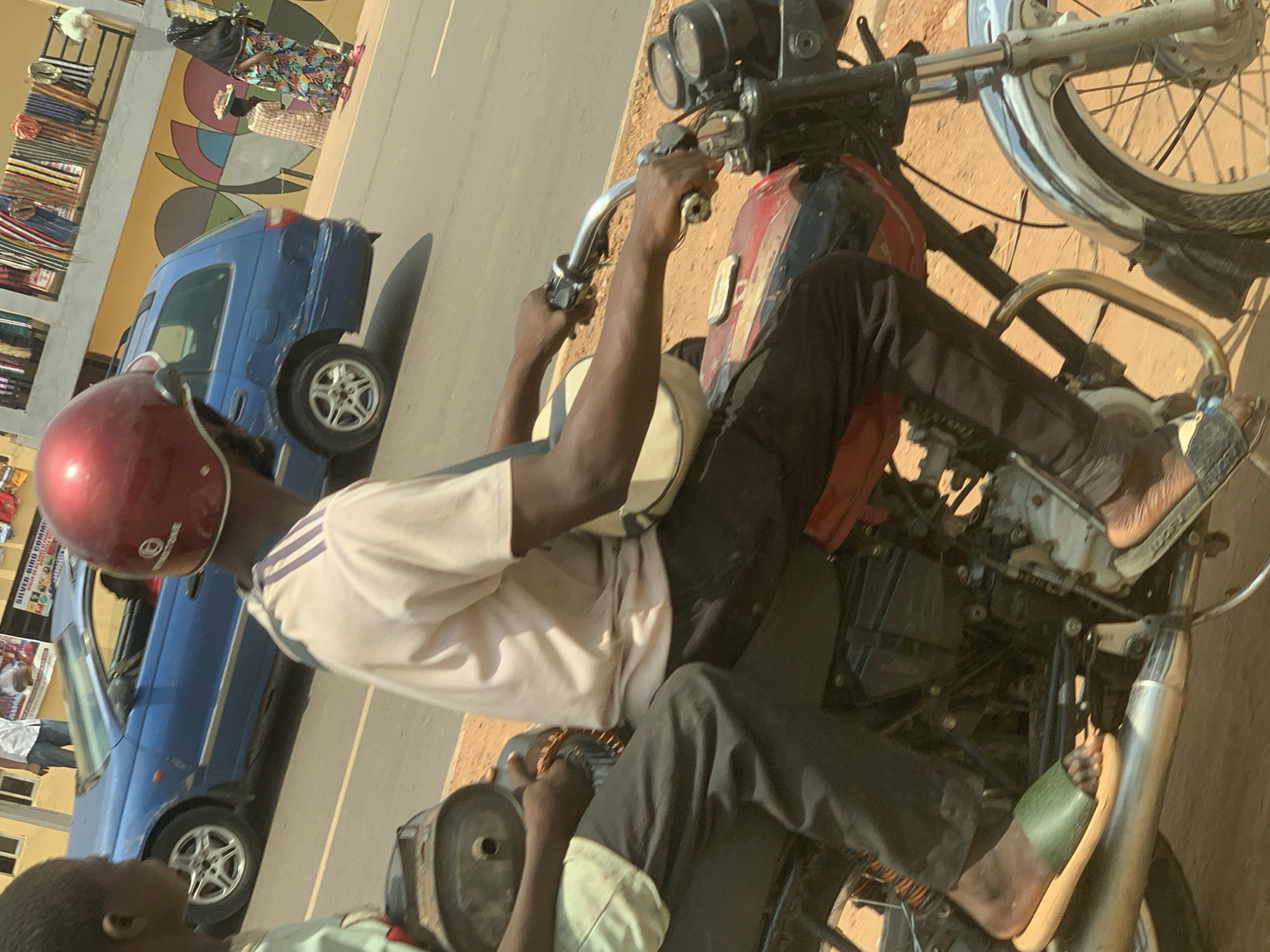 A motorcycle is parked on the side of a street, oriented with the front wheel pointing towards the right edge of the frame. A man wearing a maroon helmet, a light-colored shirt with dark stripes on the sleeves, and dark pants sits on the motorcycle. His hands are on the handlebars. Another individual, partially visible in the lower left corner, is wearing a light-colored shirt. A blue car is parked behind the motorcycle, oriented in the same direction. In the background, a building with merchandise displayed and a large, colorful mural can be seen. The street is paved, and the scene appears to be within a residential or commercial area of Ado, Nigeria.