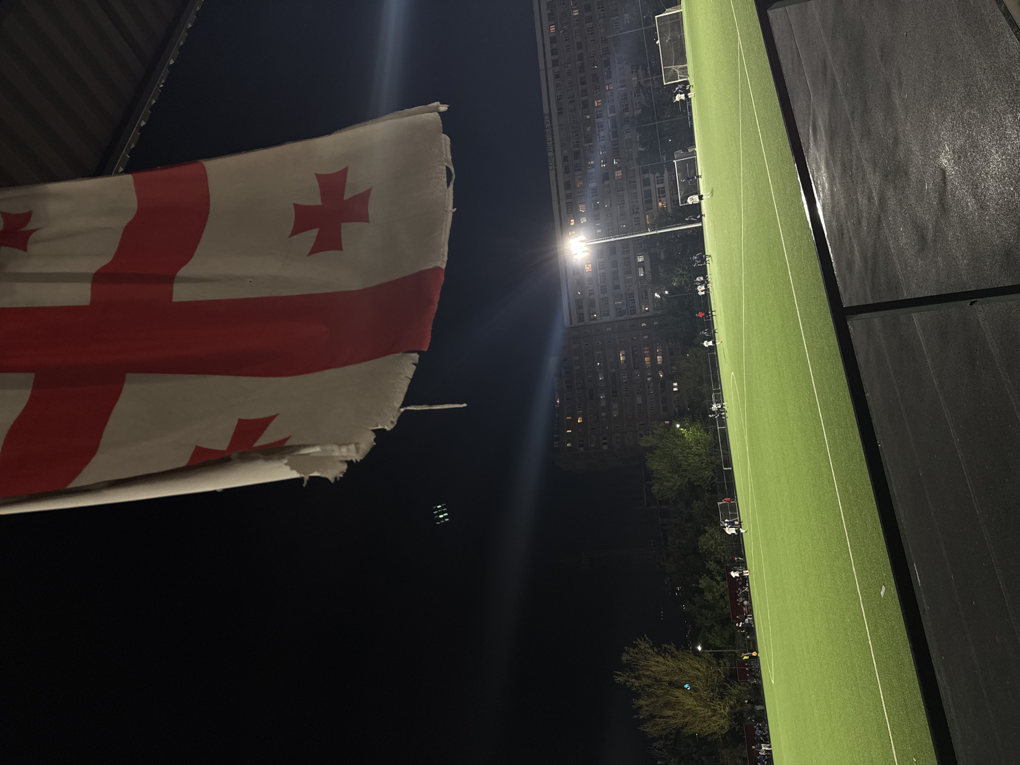 The image captures a nighttime scene featuring a prominent Georgian flag in the foreground, overlooking a brightly lit football field. The flag, white with a large red St. George's cross and four smaller Bolnisi crosses in each quadrant, appears worn and frayed along its edges.
Below and to the right of the flag (when oriented correctly), a green artificial turf football pitch is visible, marked with white lines and featuring goals at both ends. Several individuals, likely players, are scattered across the field, suggesting a match or practice is underway or has recently concluded. The field is intensely illuminated by floodlights, creating a strong glare in the upper portion of the frame.
In the background, a large, multi-story residential or apartment building stands tall, its numerous windows casting light into the dark night, further confirming the time of day. Trees line the perimeter between the field and the building. Along the right side of the field, a dark surface, possibly a running track or spectator area, is visible. Faint text "LIVE LIFE FOREVER" can be discerned on the side of the background building. Given the flag and provided context, the scene is set in Tbilisi, Georgia. The photograph is taken from an elevated vantage point, possibly from a stand or adjacent structure.