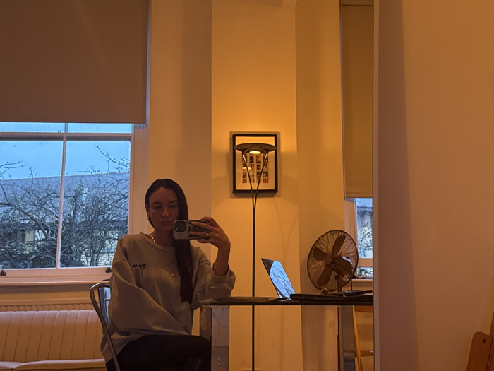 The image is a mirror selfie taken indoors, depicting a female subject in an apartment or room in London, United Kingdom, at dusk or evening. A woman with long dark hair, wearing a light grey sweatshirt and dark trousers, is seated, holding a smartphone in her right hand to capture her reflection. She wears a gold-colored necklace and rings on her left hand. In front of her is a dark-surfaced table with metallic legs. An open laptop with an illuminated screen is positioned on the table to the left, and a vintage-style bronze desk fan is situated to the right. Behind the table, two tall sash windows are visible. The left window features a beige roller blind partially lowered; the right window's blind is fully raised, revealing bare tree branches and the roofs of residential buildings, consistent with London architecture, under a blue twilight sky. Between the windows, against a warm, light-colored wall, stands a dark floor lamp. A framed artwork, possibly abstract with colorful elements, hangs on the wall behind the lamp. A light-colored, ribbed sofa or bench is partially visible to the left.