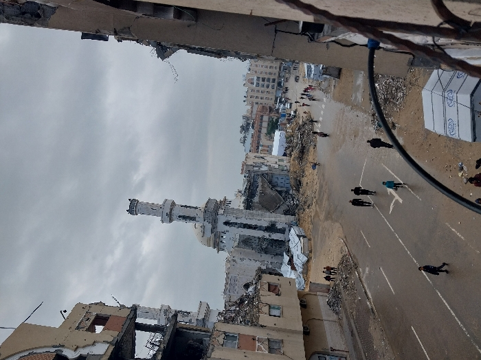 An elevated view captures a scene of extensive urban destruction in Khan Yunis, Palestinian Territory, under an overcast sky. In the center-left, the heavily damaged Great Mosque of Khan Yunis (Al-Kabir Mosque) stands with its white minaret largely intact but showing visible structural damage, while the main mosque building behind it is largely reduced to rubble and skeletal remains. Multi-story residential or commercial buildings on both the left and right sides of the frame are severely damaged, with facades collapsed, exposing interior spaces and twisted rebar.
A wet, dark-paved road with faded white markings runs through the midground, occupied by approximately a dozen individuals walking. Most are moving away from the camera; five people are visible in the immediate midground, and a larger group is further down the street towards the right, some appearing to walk in a line. The paved road transitions into a vast expanse of dirt and building debris, indicative of widespread destruction across the urban landscape. In the far background, additional damaged structures and some white tent-like shelters are discernible. A dark, curved metal railing from the vantage point building partially obstructs the immediate foreground, and a white, rectangular structure with blue text is visible below it on the right.