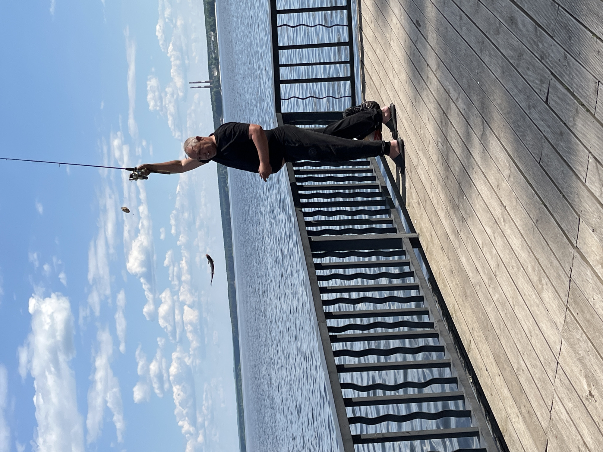 A man is fishing from a wooden pier on a sunny day. He is wearing a black t-shirt and black pants and is holding a fishing rod. A fish is airborne to the left of the man. The pier is next to a large body of water, possibly the Kama River or a reservoir. In the background, there are trees and two industrial smokestacks. The sky is blue with white clouds.