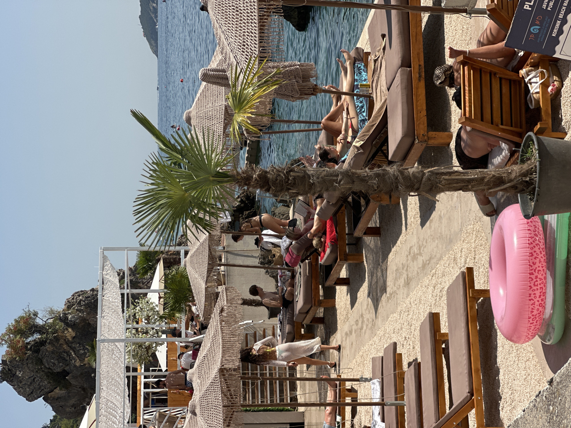 This is a photograph of a beachside cafe or club in Budva, Montenegro, with the Adriatic Sea in the background. Several people are relaxing on lounge chairs under thatched umbrellas. A woman in a white cover-up walks on the gravel beach, while others are seated or lying down. The scene includes palm trees, lounge chairs, umbrellas, and the sea. A large rock formation with vegetation is visible on the left side of the image, and hills form the horizon. A pink inflatable ring is in the foreground.