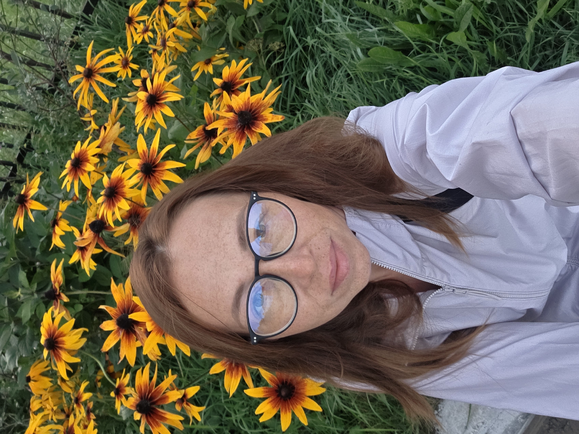 In this close-up, high-angle selfie, a woman with reddish-brown hair and freckles is positioned in the foreground. She is wearing black-rimmed glasses and a light purple zippered jacket. Behind her, a dense backdrop of vibrant yellow and orange flowers, possibly Black-Eyed Susans, creates a natural setting. The flowers are in full bloom, with dark centers and elongated petals. The background also includes green foliage and hints of a dark fence and a concrete surface. The scene appears to be outdoors, likely in a park or garden, suggesting a location within the city of Chaikovsky, Russia, given the context.