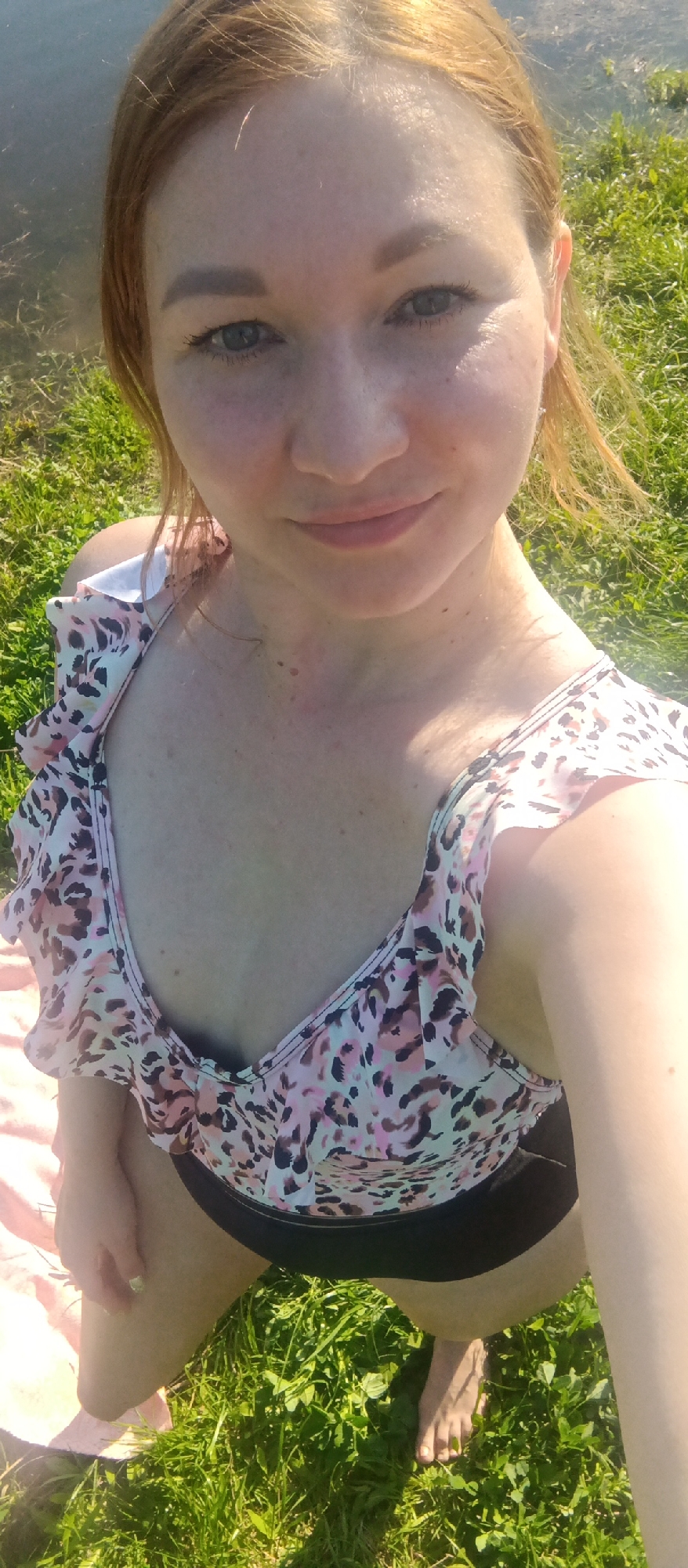 A woman with reddish-blonde hair is taking a selfie outdoors. She has fair skin, light eyes, and is wearing a swimsuit with a pink and brown animal print top and black bottoms. The swimsuit top has ruffles at the shoulders. The background includes green grass and a body of water. Part of a pink towel or blanket is also visible. The location appears to be a grassy area near water in the city of Unknown, Unknown.