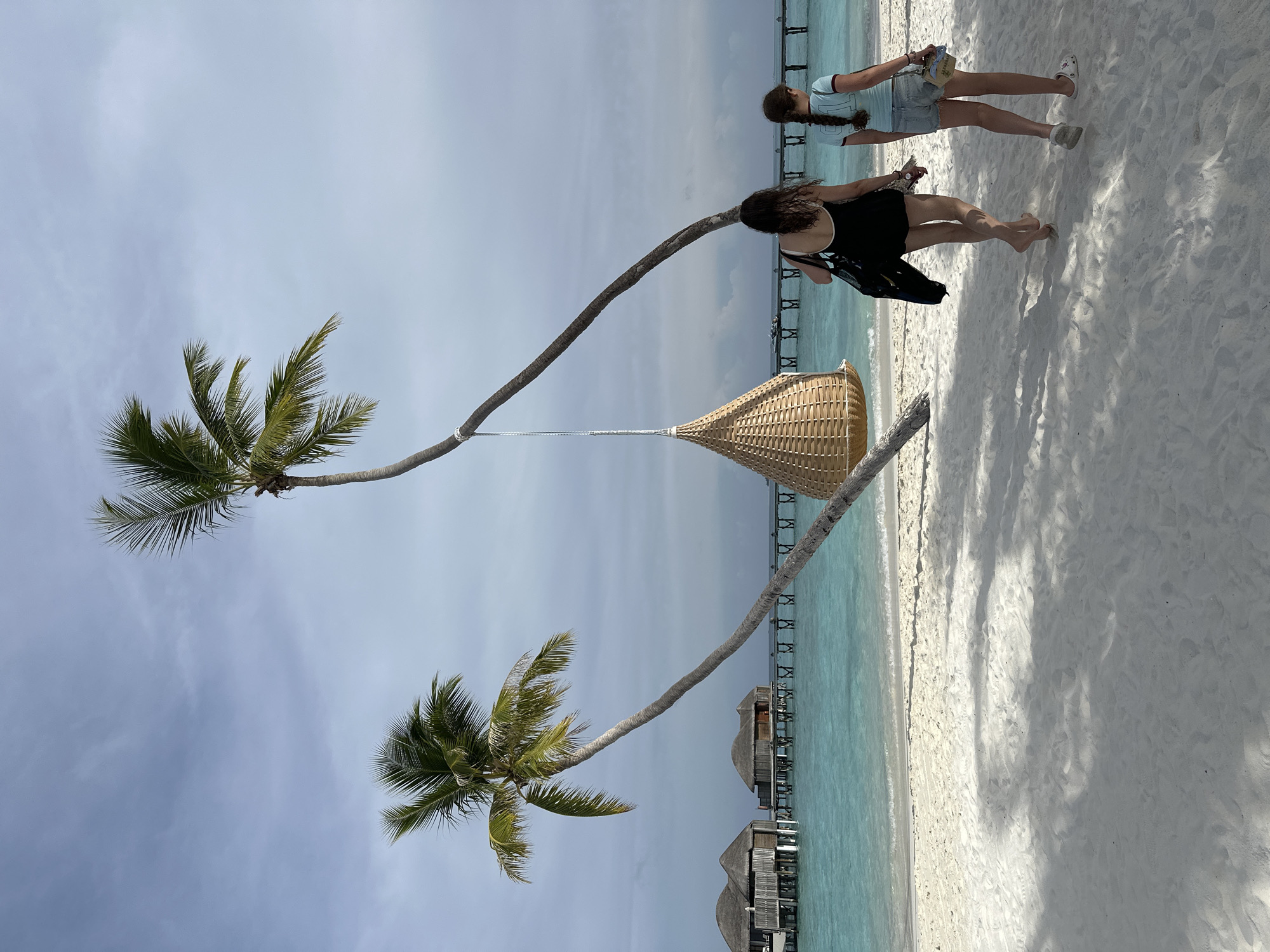 The image depicts a tropical beach scene in Halaveli, Maldives. Two individuals, a woman and a girl, are walking away from the camera on a white sandy beach towards the turquoise ocean. In the background, overwater bungalows with thatched roofs are visible, connected by a wooden pier extending into the water. Two leaning palm trees frame the scene, with a woven swing hanging from one of them. The sky is overcast with soft clouds. The woman is wearing a black swimsuit and carrying a black bag, while the girl is in a light blue t-shirt and denim shorts, carrying a small bag. The water is clear, and small waves are lapping at the shore. The overall atmosphere is serene and idyllic.