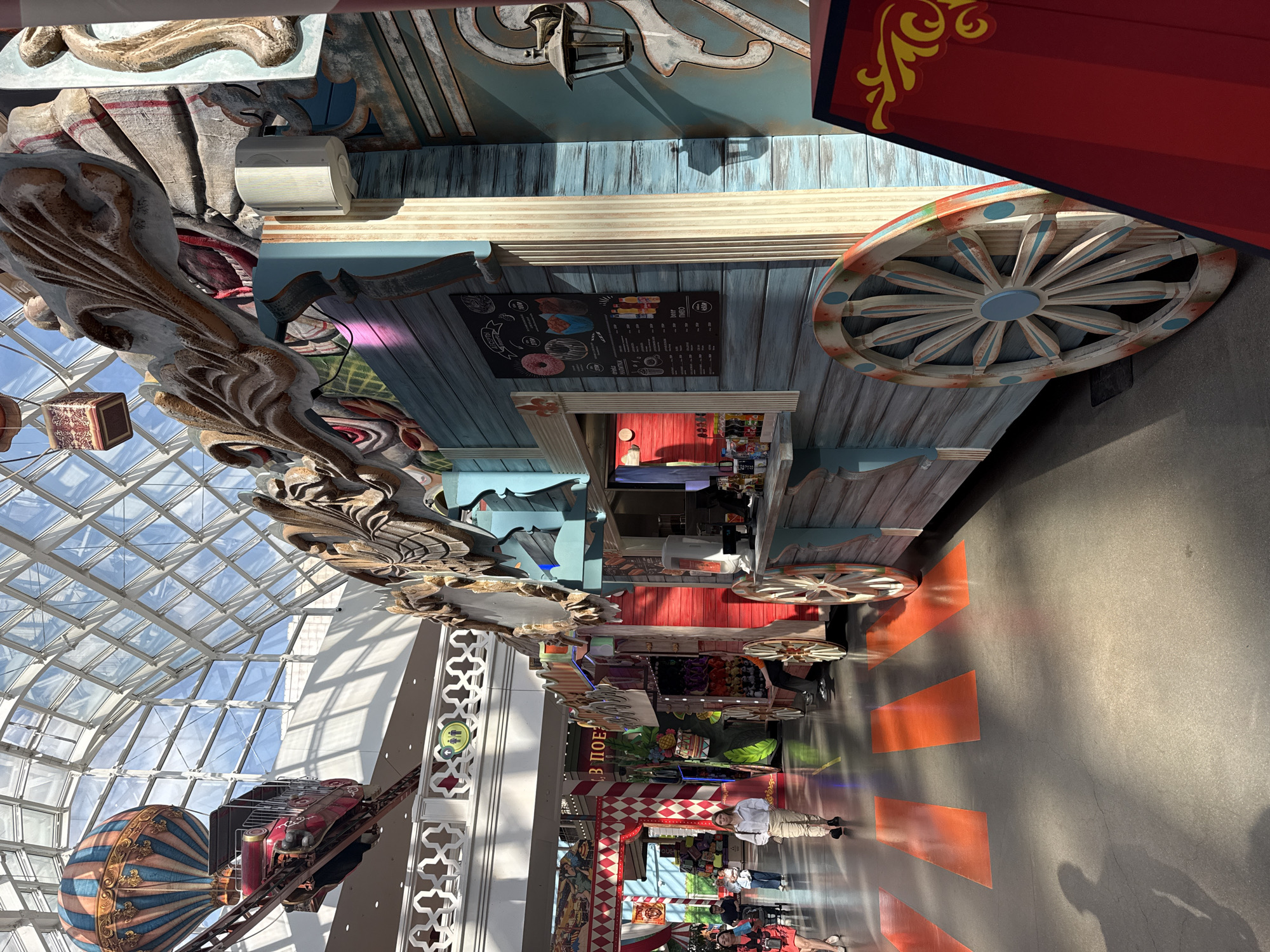 This image shows the interior of the "Dream Island" theme park, a large entertainment complex in Moscow, Russia. The location is a brightly lit indoor space featuring a glass ceiling and a decorative, ornate ceiling structure on the left.
The scene includes:
* A food stall with a wood-paneled exterior and a menu board.
* Decorative oversized wooden wheels attached to the stall.
* An air balloon roller coaster with an inflated hot air balloon.
* Several people, some of whom appear to be customers and others who are park staff.
* A pathway in the foreground marked with orange triangles, possibly to indicate a route or section within the park.
* Various other decorative elements and structures characteristic of a theme park, including an entrance with red and white harlequin pattern.