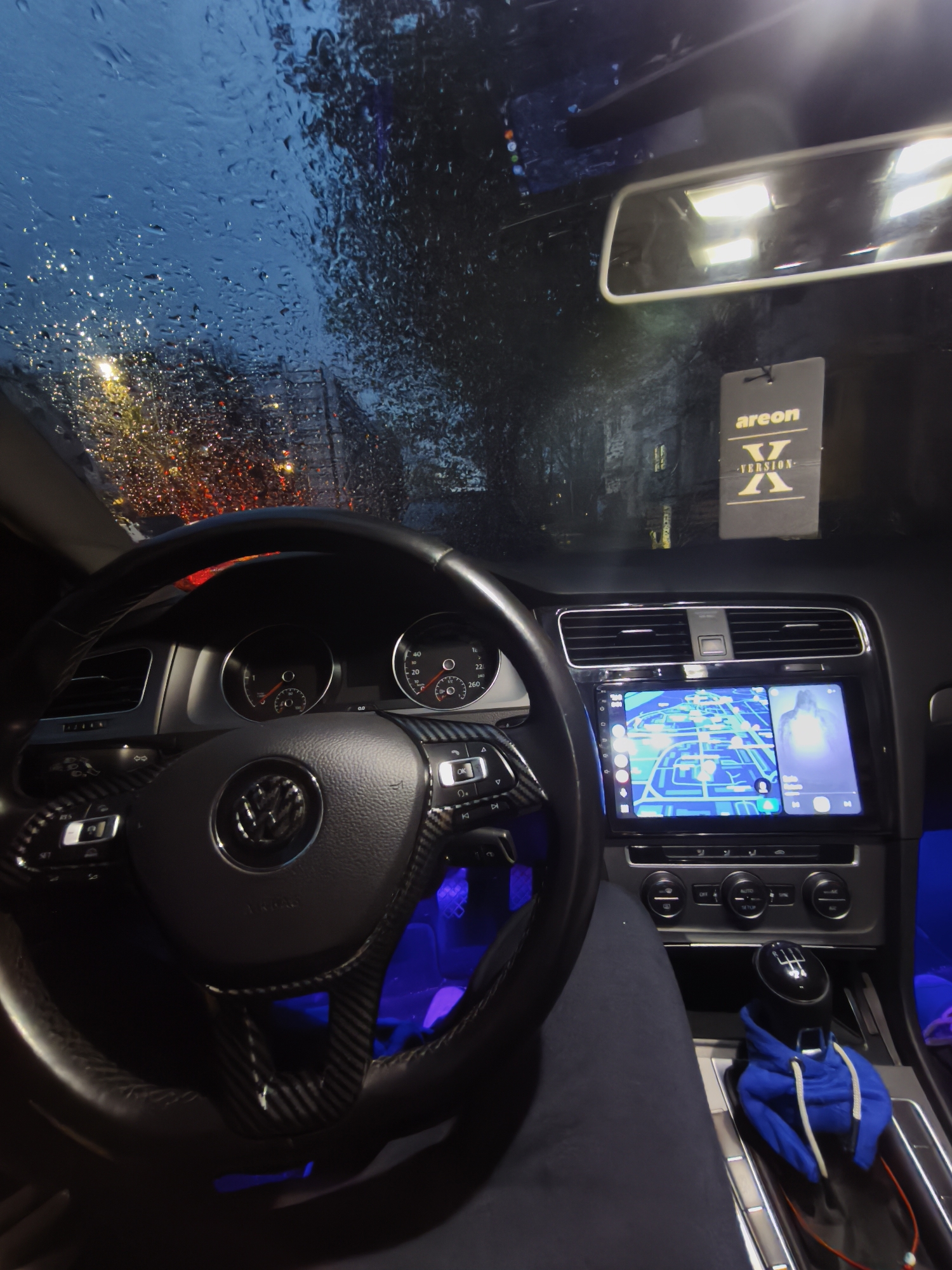 The image captures the interior of a Volkswagen vehicle, likely a Golf, at night or dusk during heavy rain in Burgas, Bulgaria. The perspective is from the driver's seat, looking forward through the windshield.
The windshield is covered in numerous raindrops, creating a blurred, abstract view of the outside world. Through the rain-streaked glass, streetlights and city lights glow in warm orange, yellow, and red hues, reflecting off the wet surfaces, contrasting with the dark blue sky and indistinct shapes of buildings.
Inside the car, a black leather steering wheel with visible carbon fiber trim and a prominent silver Volkswagen logo dominates the foreground. Controls are integrated into the steering wheel spokes. Beyond the wheel, the illuminated instrument cluster shows dials for speed and RPM. The dashboard features air vents and a large, brightly lit infotainment screen. This screen is divided, displaying a blue and white navigation map on the left side and what appears to be a media player with a glowing figure on the right. Below the screen are climate controls and other buttons.
Blue ambient lighting illuminates the footwell and parts of the dashboard, casting a cool glow. A manual 6-speed gear stick is visible in the lower right, with a small blue hoodie draped over it. Part of a leg, presumably the driver's, is seen in the lower left.
An "areon X -VERSION-" air freshener, black with white and yellow text, hangs from the rearview mirror. The rearview mirror itself reflects bright, rectangular lights from within the car's cabin. The scene conveys a moody and atmospheric setting, combining the technology and comfort of the car interior with the dynamic, rain-drenched exterior.
