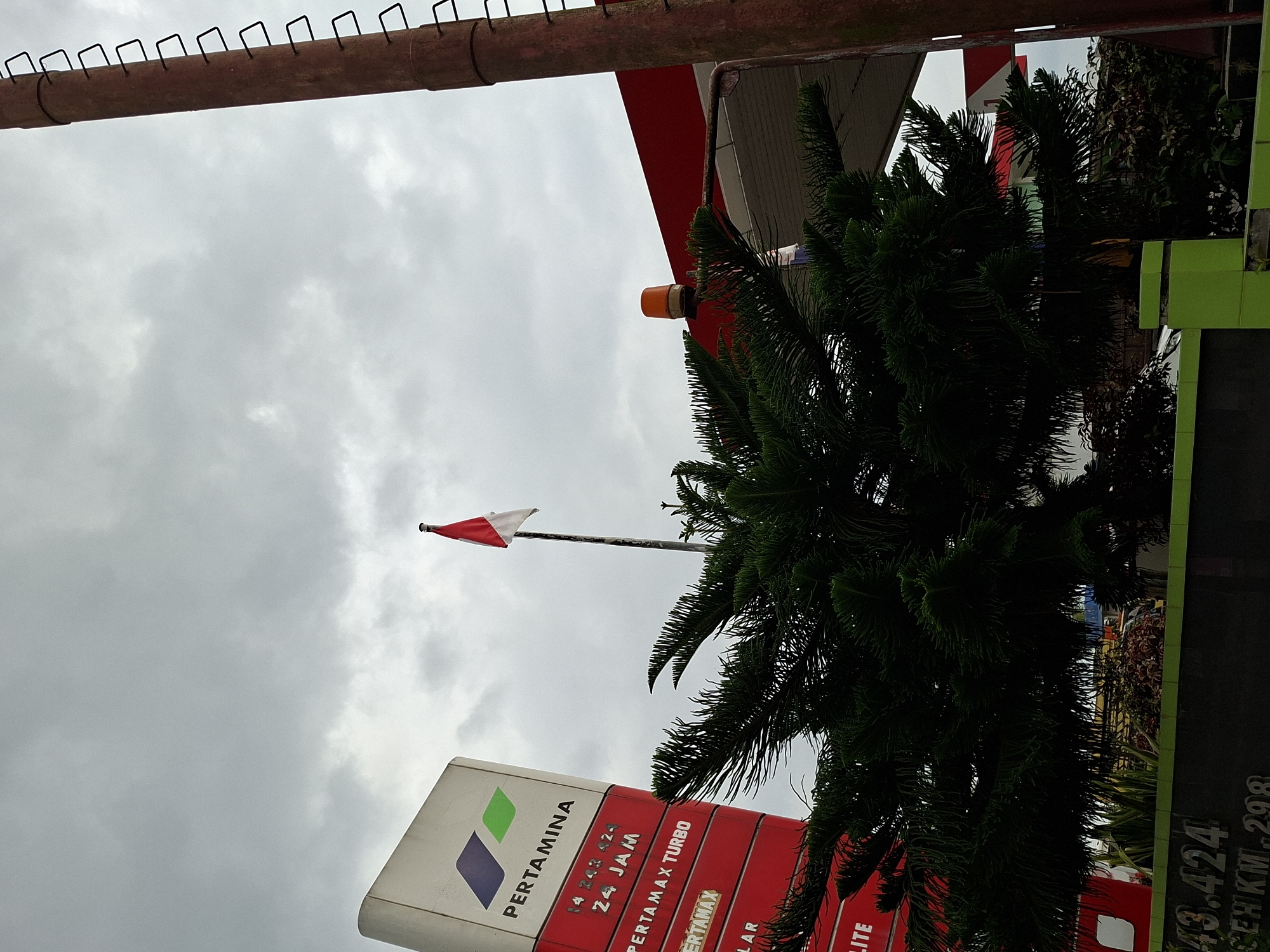The image shows a Pertamina gas station in Meunasahaluedrienlhoksukon Barat, Indonesia. A Pertamina sign is visible with the logo and text "PERTAMINA" and "PERTAMAX TURBO." The Indonesian flag is waving in the background. A pine tree partially obscures the gas station canopy. The sky is overcast. A rusted pipe structure is visible in the upper portion of the frame. A black sign with text "43.424 PERKM-298" is on the right side of the image.