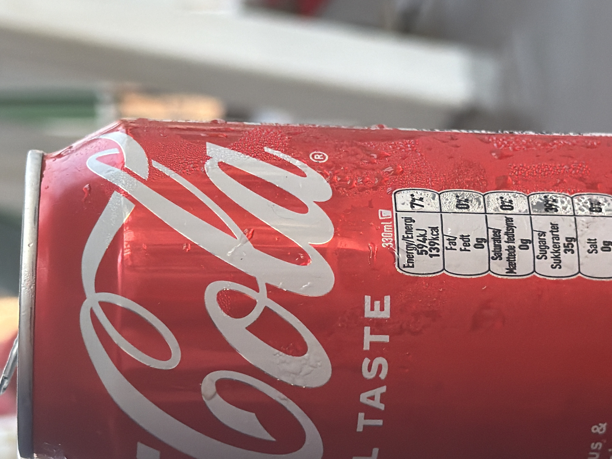 This is a close-up, angled shot of a condensation-covered can of Coca-Cola. The iconic red can features the white Coca-Cola logo and the text "REAL TASTE" in white lettering. A nutrition label is visible on the right side of the can, detailing energy content, fat, saturates, sugars, and salt. The can appears to be in an outdoor setting, possibly on a table, with a blurred background of what could be foliage or other outdoor elements. The condensation on the can suggests it is a cold beverage on a warm day. The overall impression is one of refreshment and enjoyment of a popular soft drink.
