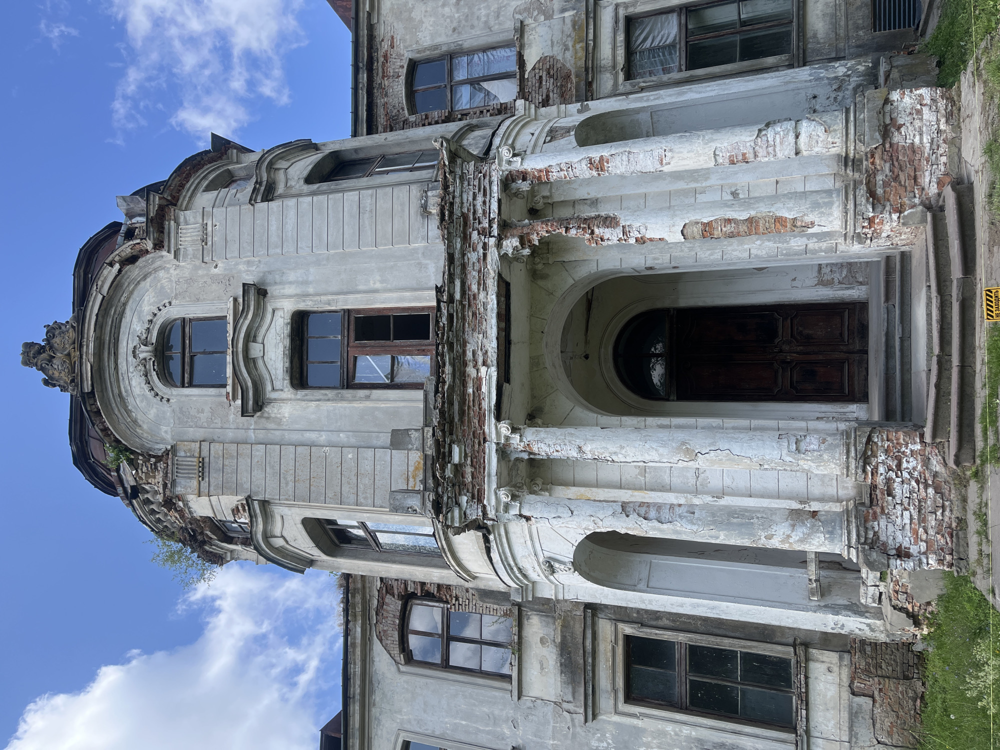 This is a low-angle, full shot of the dilapidated facade of a classical-style building, likely a manor or palace, under a partly cloudy blue sky. The structure is composed of pale, weathered stucco over brick, with significant signs of decay. The central section features a portico with arched entrances and damaged white columns, revealing exposed red brickwork. Above the portico is a damaged balcony with crumbling brick and plaster. The upper floors have tall, arched windows, many with broken or missing panes, some covered with translucent material. Decorative architectural elements are visible at the roofline, where vegetation is growing from cracks. The surrounding grounds appear overgrown with grass. A yellow warning sign is partially visible in the foreground. The building's architectural style suggests a historical, possibly 19th-century, origin. The overall impression is one of abandonment and decay.