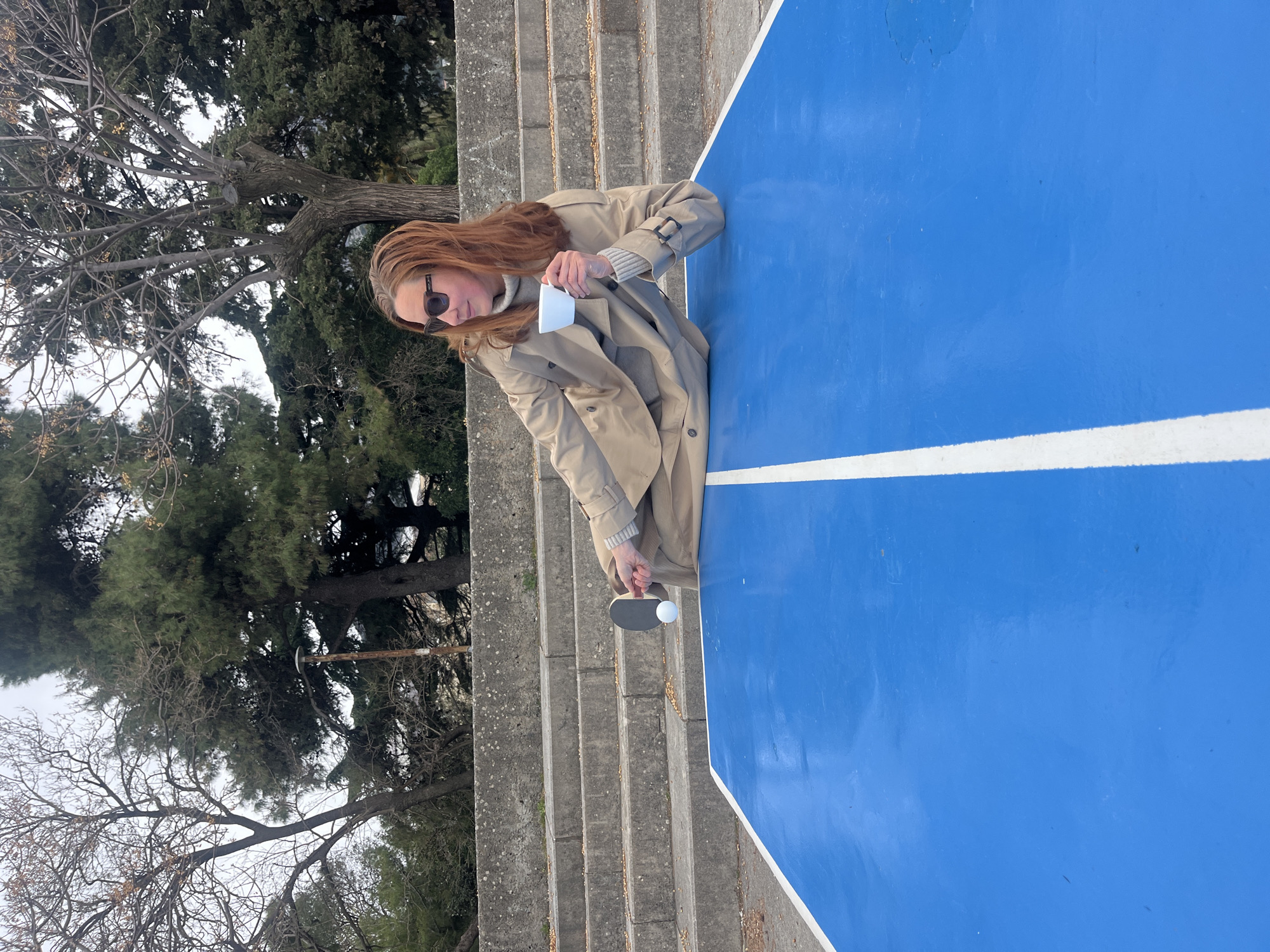 A young woman with long, reddish-brown hair, wearing a tan trench coat, sunglasses, and a light-colored sweater, is standing on concrete stairs next to a bright blue ping pong table with a white stripe. She's holding a ping pong paddle in her right hand, with a ping pong ball next to it. In her left hand, she holds a white, rectangular object, potentially a phone or card. The scene appears to be outdoors, with trees visible in the background and the stairs leading to a higher level. The ping pong table is likely located in a public space in the city of Budva, Montenegro, since this is where the images were shot. The table and stairs suggest an area for recreation or leisure. The woman is engaged in the activity of preparing to play ping pong.