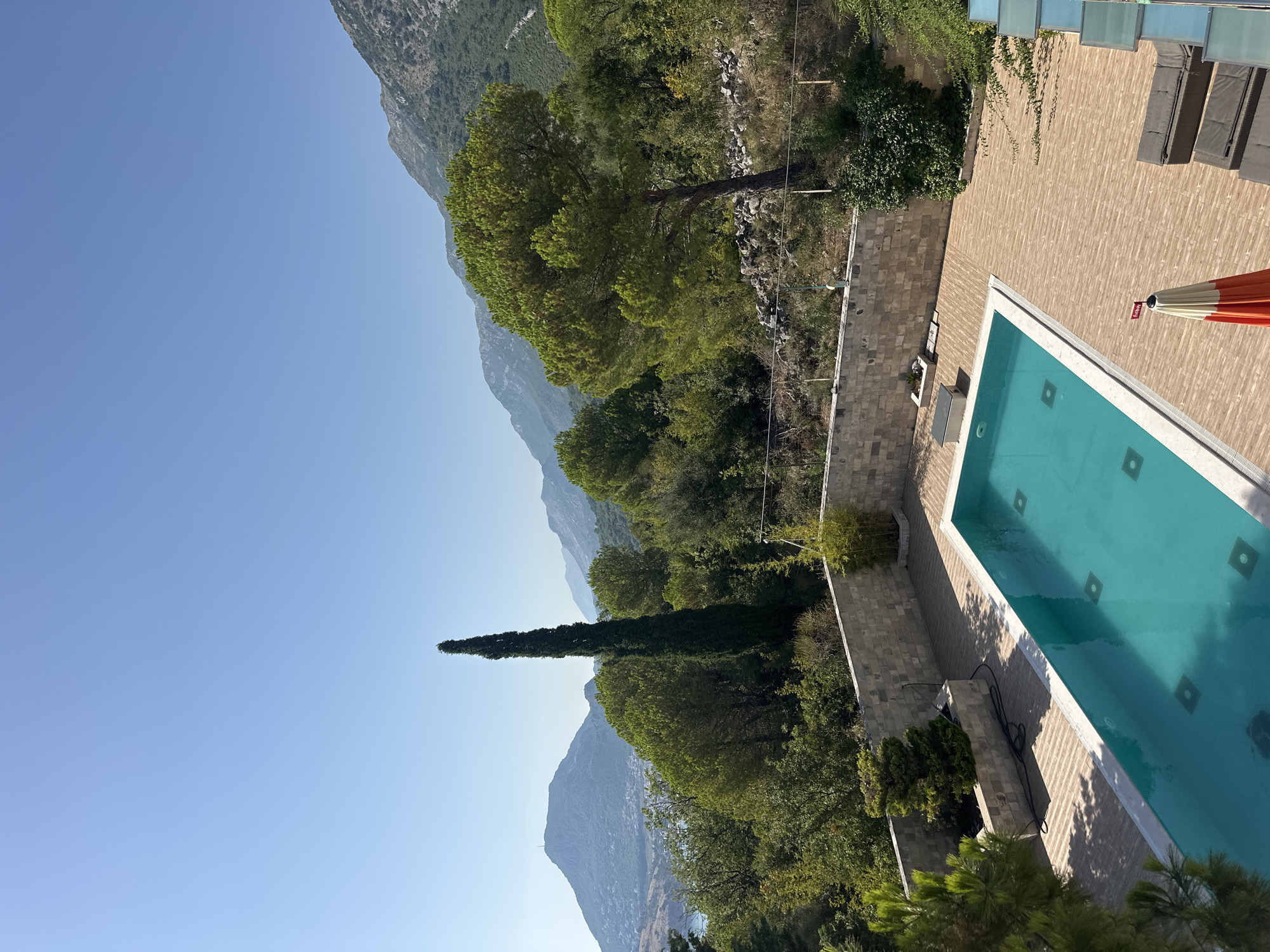 This is an eye-level, medium shot captured on a sunny day in Brca, Montenegro, showcasing a swimming pool area with a scenic mountainous backdrop. The foreground is dominated by a rectangular swimming pool filled with clear turquoise water. The pool's edge is lined with white tiles, and its bottom features a pattern of square, dark-colored indentations. The pool is surrounded by a light brown wooden deck. To the right of the pool, two dark grey lounge chairs are visible, with an orange and white umbrella standing next to them, displaying a small red flag. Behind the pool, a stone wall with a rough-hewn texture encloses the area. Beyond the wall, lush green trees and vegetation cover the landscape, interspersed with rocky terrain. In the middle ground, a tall, slender cypress tree stands out prominently amidst the dense foliage, to its right is a large pine tree. The background is characterized by rolling hills and mountains, fading into a hazy blue under a clear, light blue sky. A body of water and distant hills are visible to the left, suggesting proximity to the coast. The overall scene depicts a tranquil outdoor space, likely part of a private residence or resort, enjoying a picturesque view of the Montenegrin landscape. No people are present in the frame.
