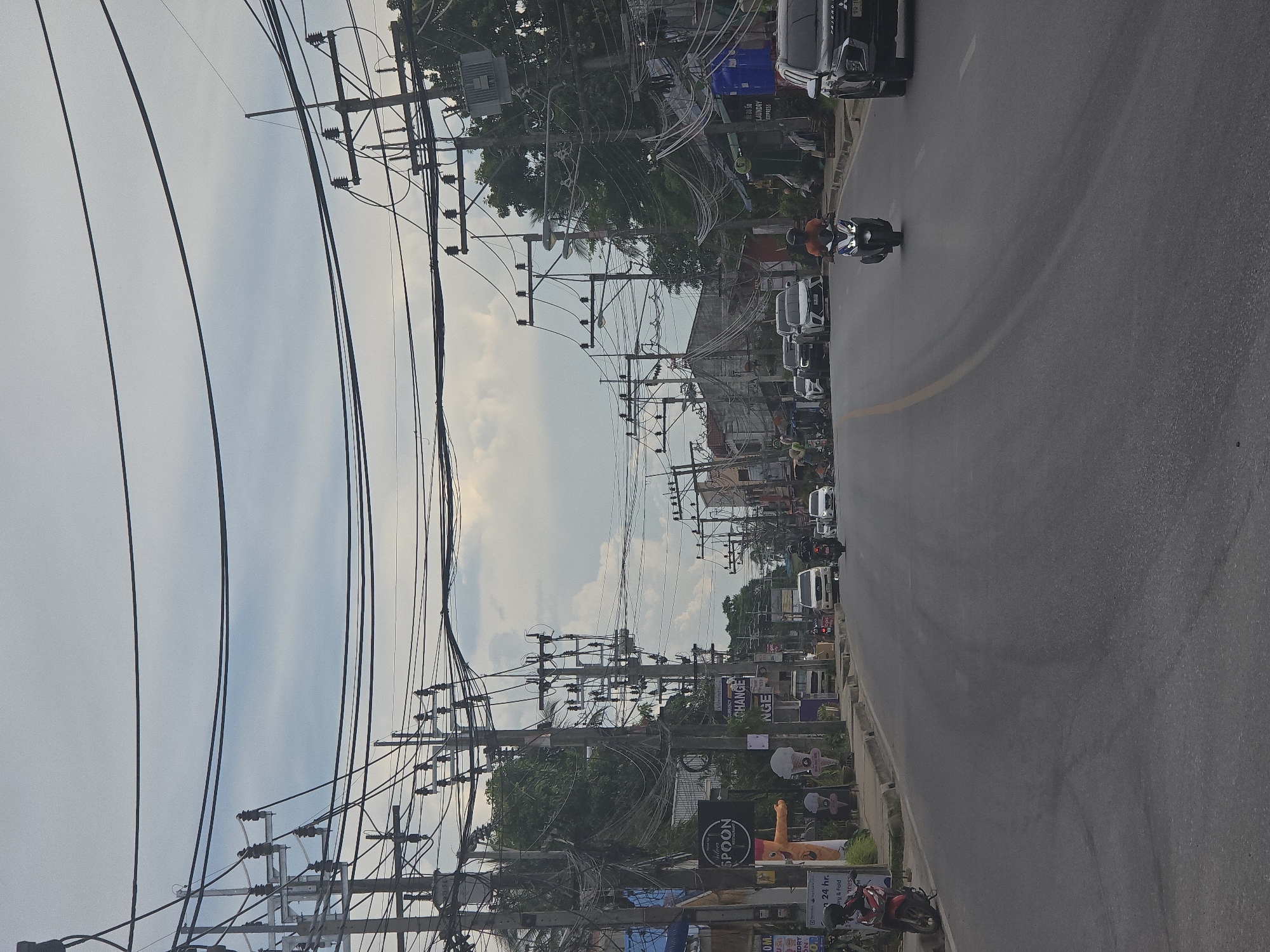 The image shows a street scene in a city, possibly in Southeast Asia, with numerous overhead electrical wires and utility poles dominating the sky. The street is paved and appears to be a two-lane road with traffic moving in both directions.
On the left side of the street, there is a sidewalk with various signs, including one for "SPOON" and another with Thai lettering. A red motorcycle is parked on the sidewalk. Further down the street on the left, there are buildings with storefronts, including what appears to be a laundry service.
On the right side of the street, there are also buildings and storefronts, with more utility poles and electrical wires. A dark-colored SUV is driving on the right side of the road, and a person on a scooter is in front of it. Other vehicles, including cars and motorcycles, are visible further down the street in both directions.
The sky is overcast with some patches of blue sky visible. The overall impression is a busy urban environment with a significant amount of electrical infrastructure.