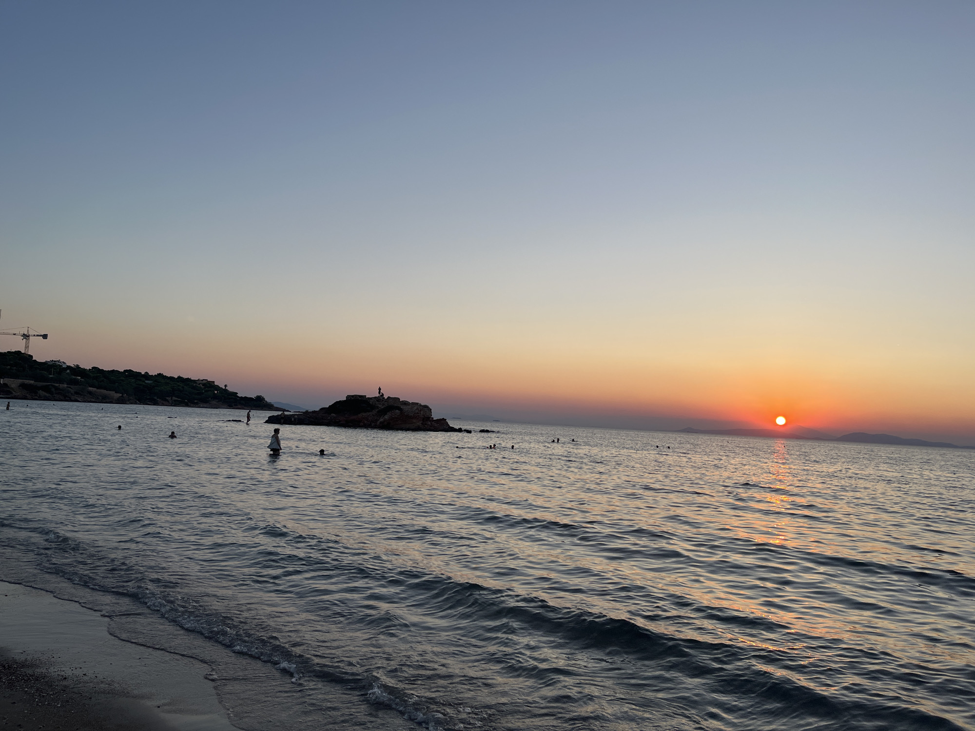 The image depicts a serene beach scene at sunset in the Vouliagmeni Municipal Unit, Greece. The sun, a bright orange orb, is low on the horizon, casting a golden glow on the water and the sky. The sky transitions from a clear pale blue at the top to a gradient of orange and pink hues near the horizon. Small waves gently lap the shore, with ripples catching the sunlight.
Several people are visible in the water. Some are swimming or wading, while others are standing near a rocky outcrop in the middle ground. On the rocky outcrop, at least two figures appear to be standing. The beach itself is dark and sandy, with the water's edge clearly defined. In the far distance, a line of hills or mountains is silhouetted against the colorful sky. A construction crane is visible on the left side of the image, suggesting some ongoing development near the coastline. The overall atmosphere is peaceful and tranquil, indicative of a late summer evening.