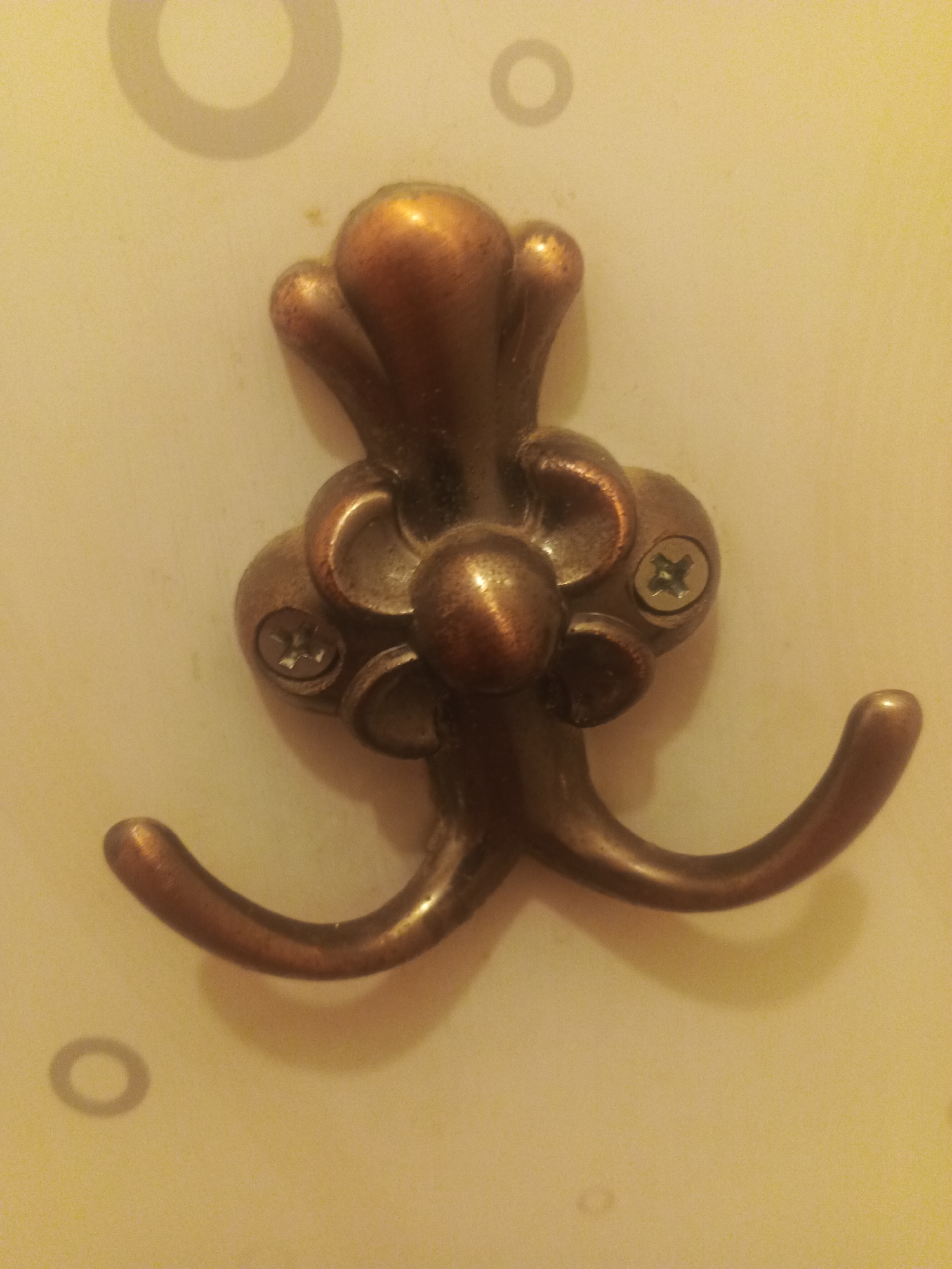 The image shows a close-up of a decorative, bronze-colored coat hook attached to a cream-colored wall. The hook has a stylized design resembling a flower or an ornate flourish, with two curved arms for hanging items. Two silver screws are visible, securing the hook to the wall. The wall has faint circular patterns imprinted on it, possibly from wallpaper or a decorative texture. The lighting casts a slight shadow from the hook. There are no people or animals present in the image. The location of this image in Biysk, Russia, cannot be determined from the visual information provided.