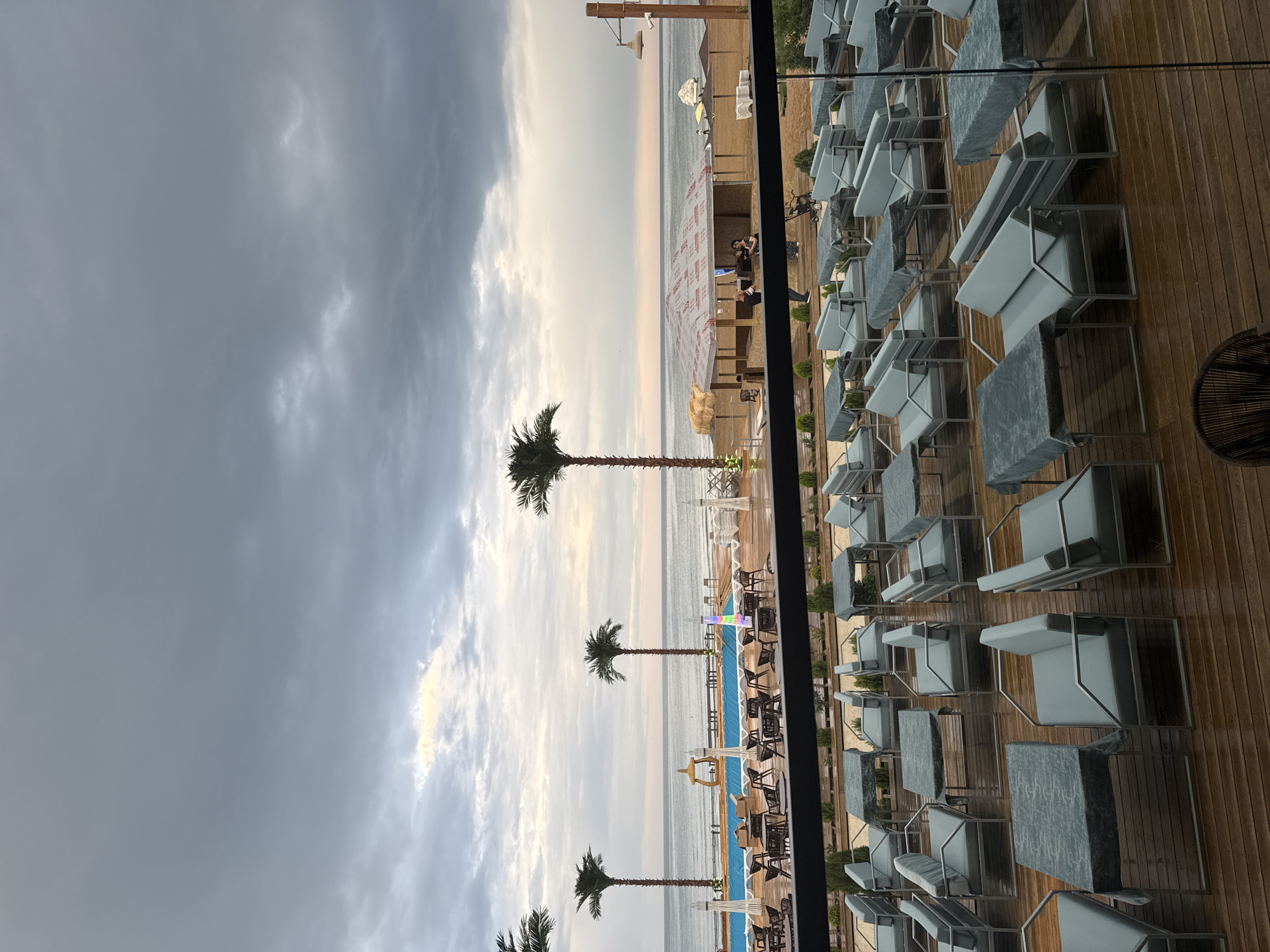 The image depicts an outdoor seating area with a view of the sea, likely a resort or restaurant. The scene is oriented vertically, with the sky and sea occupying the left portion and a wooden deck with numerous lounge chairs and tables filling the right. Several palm trees are scattered across the landscape, some closer to the foreground and others further back near the water. The sky is overcast with dramatic clouds, suggesting either early morning or late afternoon, with a hint of light breaking through on the horizon. The sea is calm, stretching out to meet the sky. The seating area features modern, light-colored lounge chairs and tables with stone-patterned surfaces, arranged neatly on a wooden deck. Some of these chairs are covered, indicating they might be unoccupied or protected from the elements. In the middle ground, there's a glimpse of a swimming pool and a beach area with umbrellas and some people visible. The overall atmosphere is tranquil, with a touch of luxury suggested by the well-maintained seating. The location is Derbent, Russia. No visible text is present in the image.