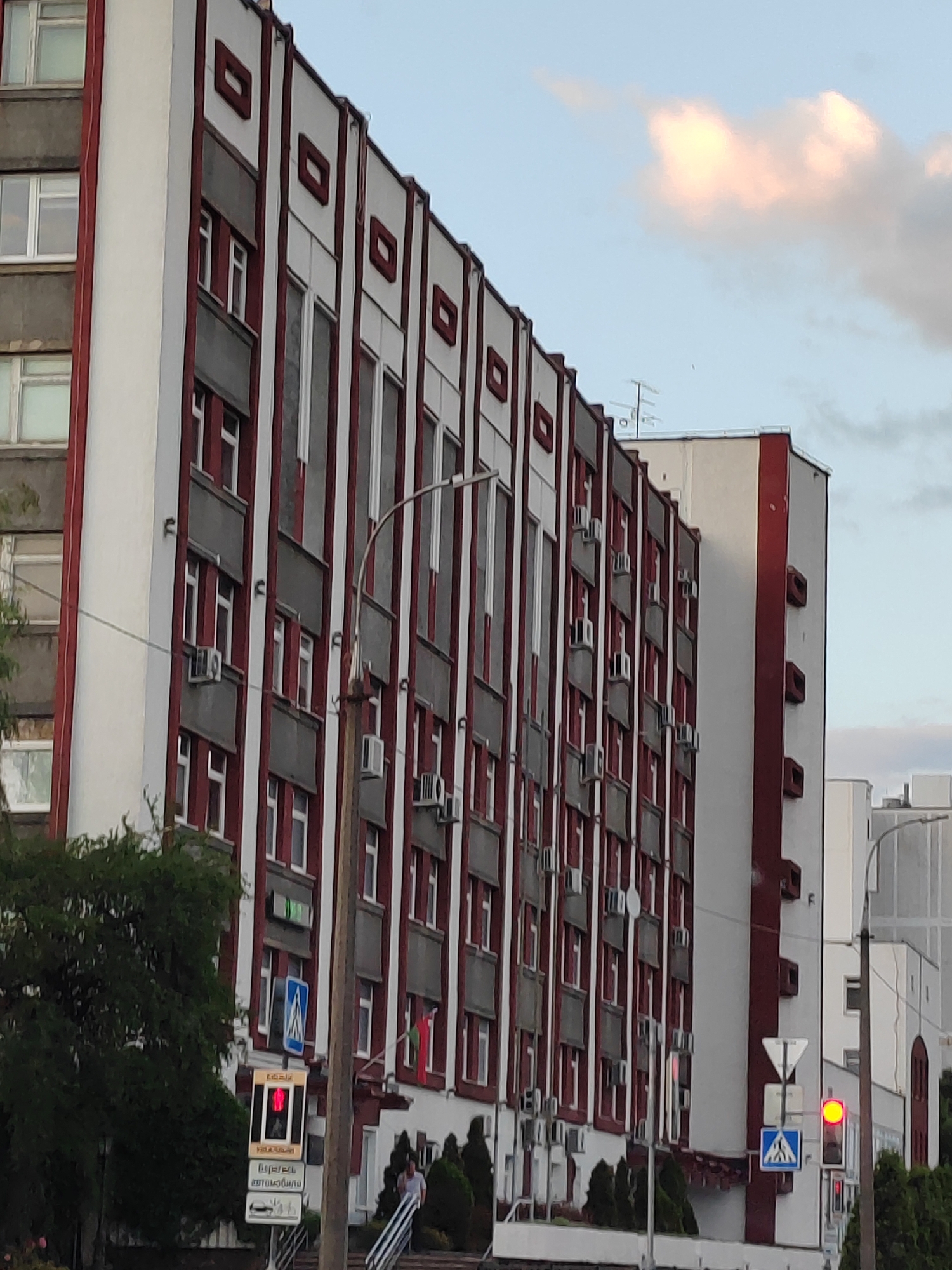 This image captures a view of a multi-story building in the city of Brest, Belarus. The building features a facade with vertical red and white striped elements, interspersed with grey sections. Numerous windows are visible, many equipped with air conditioning units. A traffic light is present in the foreground, showing a red signal. Adjacent to the traffic light is a pedestrian crossing sign. The sky above is a pale blue with a few scattered clouds, one of which is tinged with pink from the setting sun. Trees and greenery are visible along the bottom of the image, and in the background, other buildings can be seen, suggesting an urban environment.