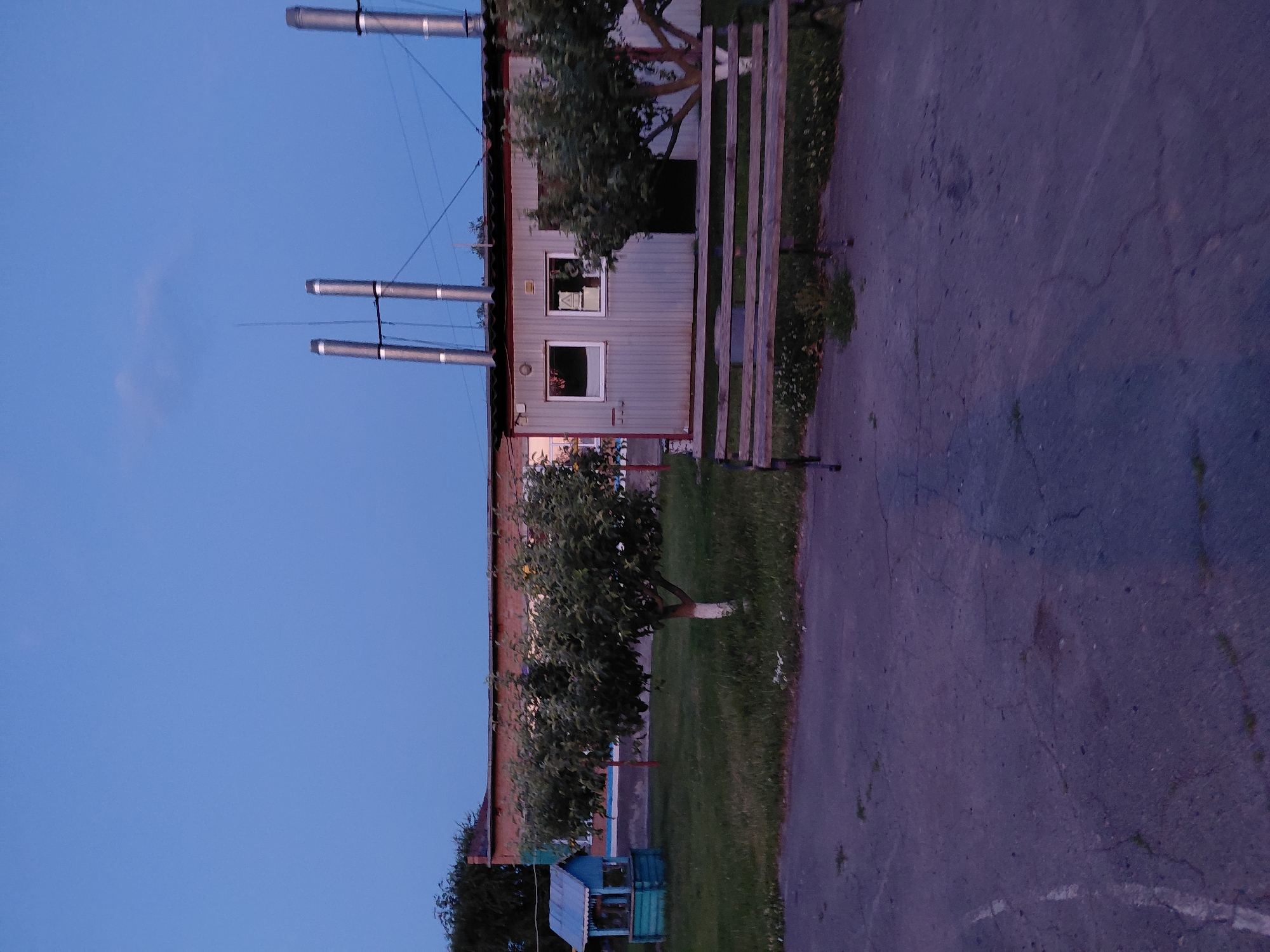 The image depicts an outdoor scene at dusk in the city of Ustyvytsia, Ukraine. In the background, a long, single-story brick building with a dark roof is visible. Attached to this building is a smaller, grey, corrugated metal structure with two windows. Each window is illuminated from within, and the one on the right displays a triangular symbol. Two large, silver smokestacks rise vertically from the roof of the metal structure. To the left of the building, a small, blue and green wooden structure resembling a well or gazebo stands on a grassy area. In front of the metal building and smokestacks, a wooden bench with a weathered appearance is positioned on a paved surface. Trees with green foliage are scattered throughout the scene, with one tree in the foreground having a white-painted trunk. The sky above is a deep, clear blue, indicating twilight. The foreground consists of a cracked asphalt surface with some patches of grass.