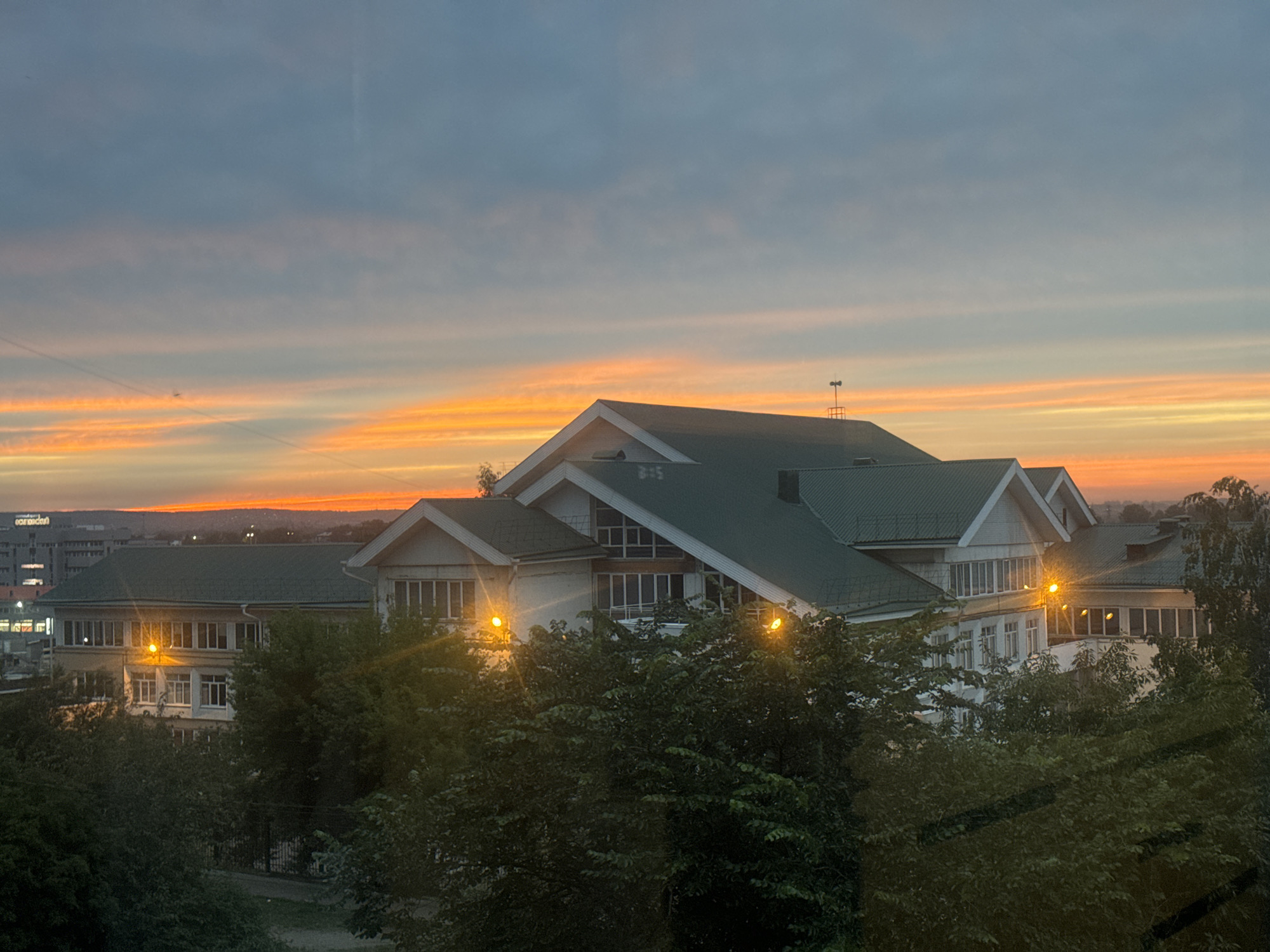 The image captures a sunset over a town, likely in Irkutsk, Russia. The sky is painted with vibrant hues of orange and yellow, transitioning into a soft blue as it rises. Wispy clouds stretch horizontally across the sky, illuminated by the setting sun.
In the foreground, several buildings with green roofs are visible, partially obscured by lush green trees. Some of these buildings appear to be educational institutions, with multiple windows and distinct architectural features. Yellow outdoor lights are on, casting a warm glow on the building facades. In the background, a cityscape with taller buildings can be seen, hinting at an urban environment.
The overall mood is serene and tranquil, characteristic of a peaceful evening. The time of day is clearly sunset, as indicated by the colors of the sky. No people are visible in the frame, and the activity appears to be simply the transition from day to night. There is no discernable text in the image.