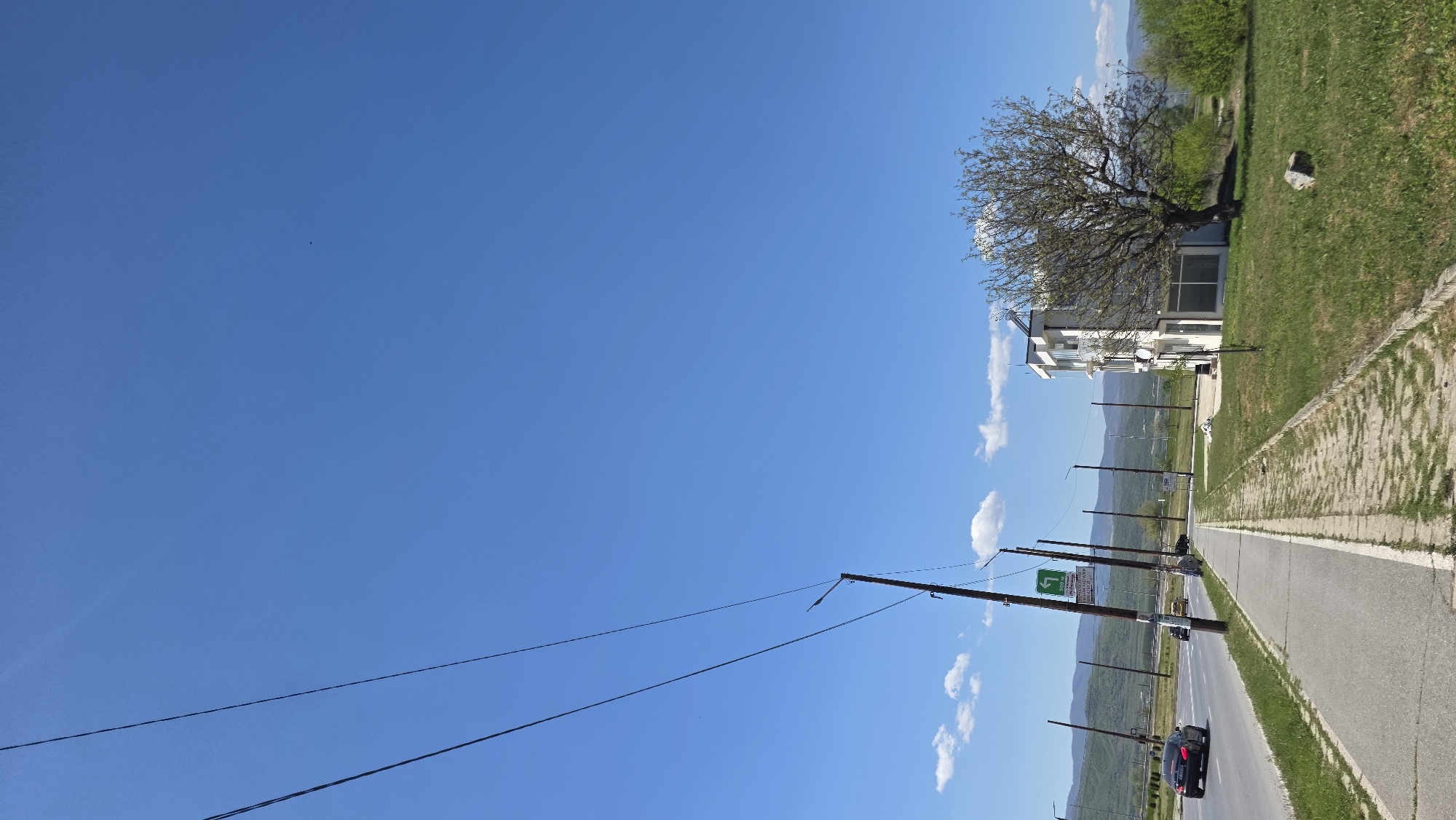 The image depicts a street scene in Blagoevgrad, Bulgaria on a clear day. The sky is blue with scattered cumulus clouds. A two-lane road with a sidewalk and grassy area runs along the right side of the frame. Utility poles line the road with wires stretching across the sky. A black car is traveling on the road. A building with a tree next to it is located next to the grassy area. Mountains are visible in the distance.