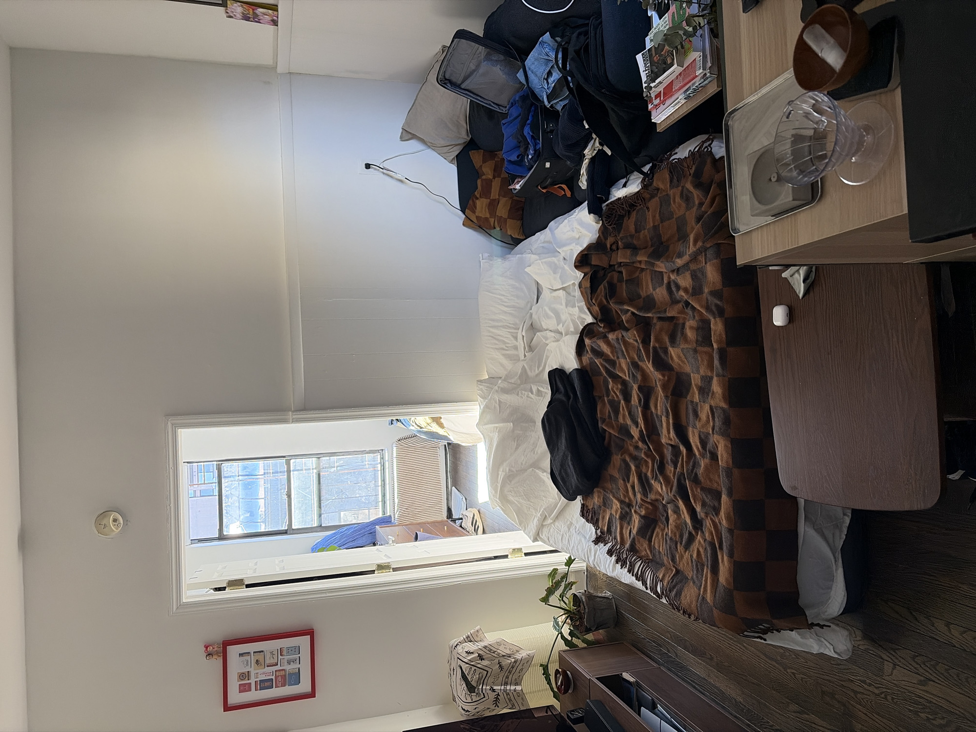 The image depicts an interior room, likely a bedroom or studio apartment, in an urban environment. The specific location within New York, United States of America, cannot be determined from the visual evidence.
The room features white walls, dark hardwood flooring, and a white ceiling with a centrally mounted smoke detector. Natural light enters through an open white doorframe on the left, revealing an adjacent room with a window secured by a metal grate, a visible radiator below it, and parts of other buildings outside. A brown dresser and light-colored athletic shoes are partially visible in the adjacent room.
The primary furniture in the main room is a bed with a white mattress, white sheets, and a brown and black checkered blanket with fringes. A crumpled black garment rests on the sheets. Adjacent to the bed on the right is a dark-colored sofa or daybed, cluttered with pillows (one light beige, one brown checkered), an open black suitcase containing blue garments, and additional clothing and bags. A white electrical outlet with a black cord is visible on the wall above the sofa. A stack of books and magazines rests on a surface nearby.
On the left wall, a red-framed picture containing a grid of small images is displayed, with two small decorative figures atop it. Below this, a brown media console holds a black electronic device, and a patterned cloth is draped nearby. A potted green plant in a grey pot is situated on the floor.
In the foreground, a dark brown wooden table holds a white Apple AirPods case and a small grey cloth. Another lighter-colored wooden table in the immediate right foreground displays a silver rectangular tray, a grey cylindrical object, a clear ribbed plastic coffee dripper, and a small brown bowl with a white object inside. A black object, possibly a laptop, is partially visible beneath these items. No people are present.