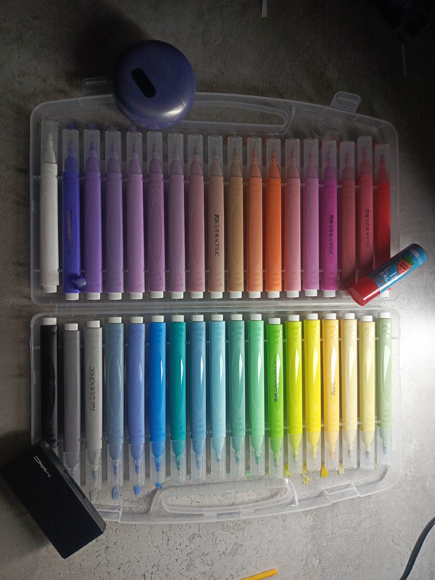 A high-angle, close-up shot shows a clear plastic case containing two rows of acrylic paint markers in a gradient of colors, ranging from white to black and including shades of purple, pink, brown, orange, red, blue, green, and yellow. A blue, round object with a black cutout is placed on top of the case. A black rectangular object with "MAC" printed on it rests near the lower left corner of the case. A tube of lip balm with a strawberry design is near the red markers. Debris is scattered around the markers. The background is a gray surface.