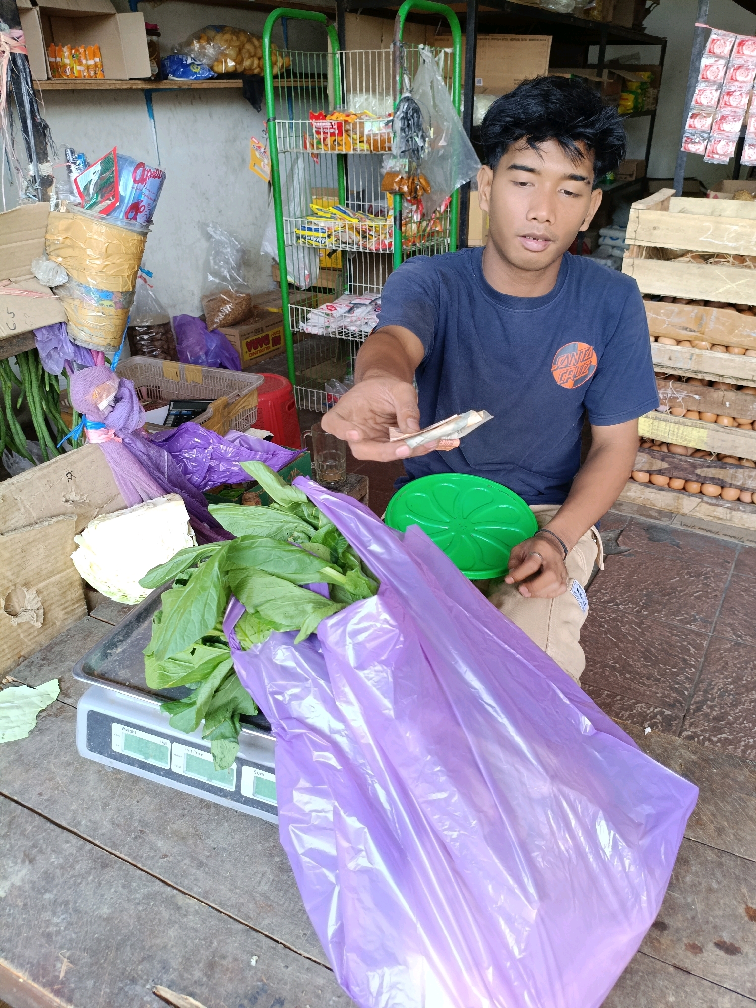 A young man, likely of Indonesian descent, is seated on a wooden surface in what appears to be an outdoor market stall in Maguwoharjo, Indonesia. He is wearing a navy blue t-shirt with a circular logo and tan pants. In his right hand, he holds a few Indonesian Rupiah banknotes, presenting them as if for a transaction. Before him, on a silver digital scale, is a bundle of green leafy vegetables wrapped in purple plastic. A bright green plastic bowl rests on his lap. In the background, shelves are stocked with various packaged goods, and wooden crates contain what appear to be eggs. The overall environment suggests a typical market setting, with goods displayed and prepared for sale.