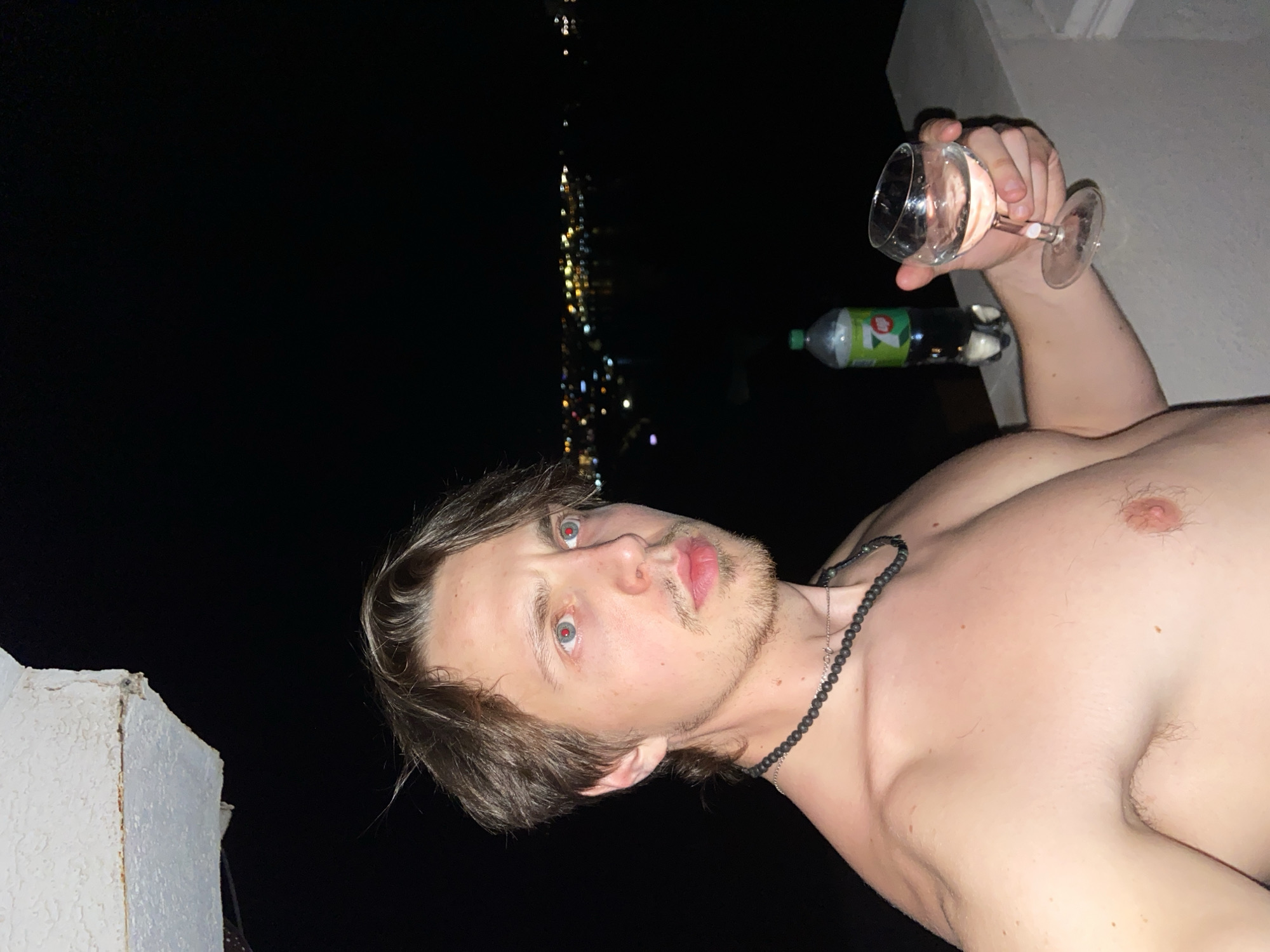 A shirtless man is holding a glass of clear liquid in his right hand and a green plastic bottle in his left hand. He has brown hair and a beard. The man is standing on a balcony with a white concrete ledge and a black sky. Lights are visible in the distance, indicating a city or settlement, identified as the city of Šušanj, Montenegro. The photo was taken at night. The man is wearing a dark beaded necklace. The image is angled upwards.