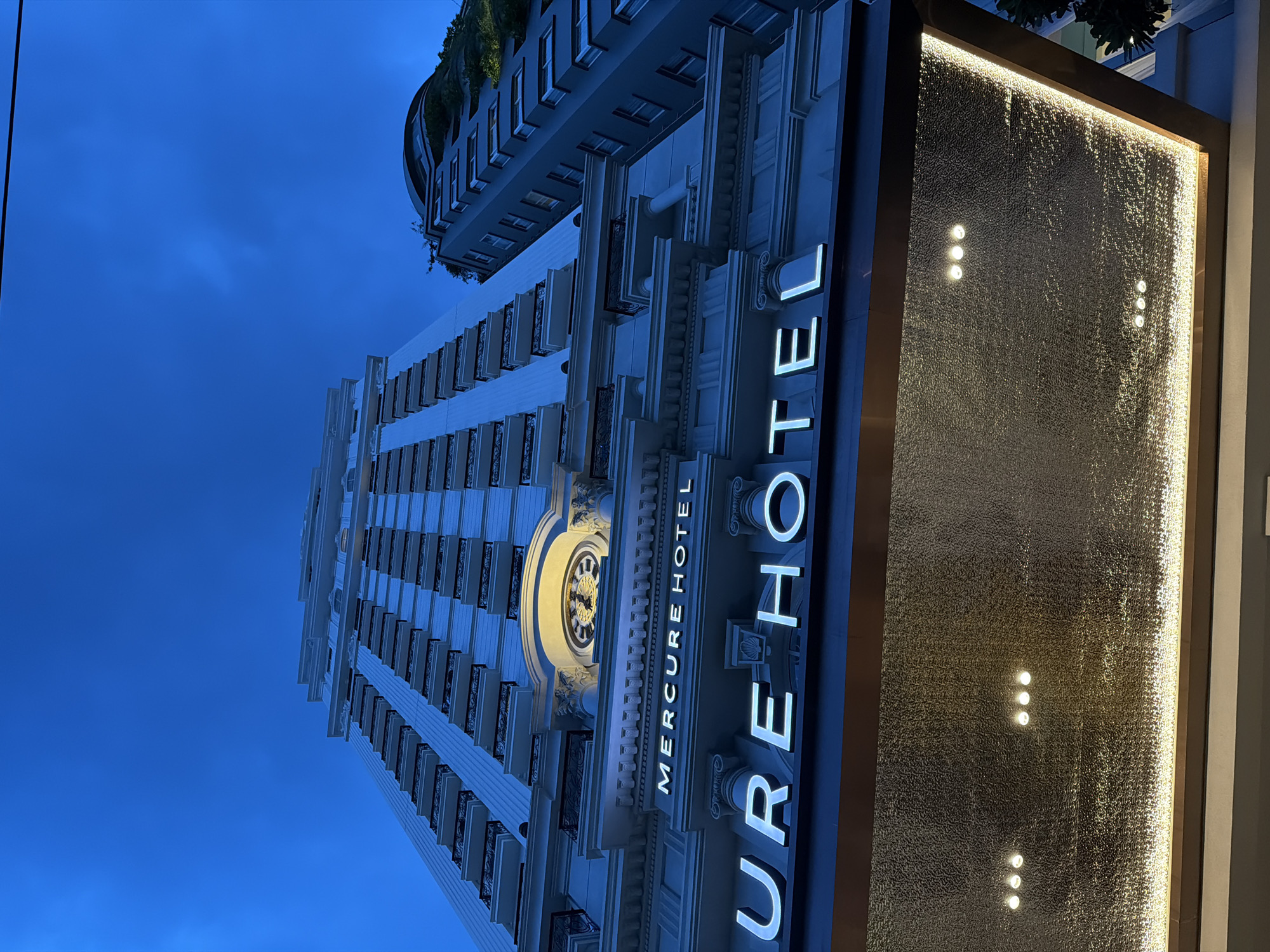 An exterior shot captures the Mercure Hotel in Nha Trang, Vietnam, at dusk. The imposing white facade of the hotel is illuminated, with rows of balconies visible on its upper floors. At the base of the building, large illuminated letters spell out "MERCURE HOTEL." Above this, a clock face is embedded in the architecture, and further up, a sign reads "MERCURE HOTEL" in smaller letters. The entrance features a textured glass ceiling with embedded lights, casting a warm glow. To the right, another building with a rounded facade and greenery on its upper levels is partially visible. The sky above is a deep blue, indicative of twilight. There are no people or vehicles present in the frame.