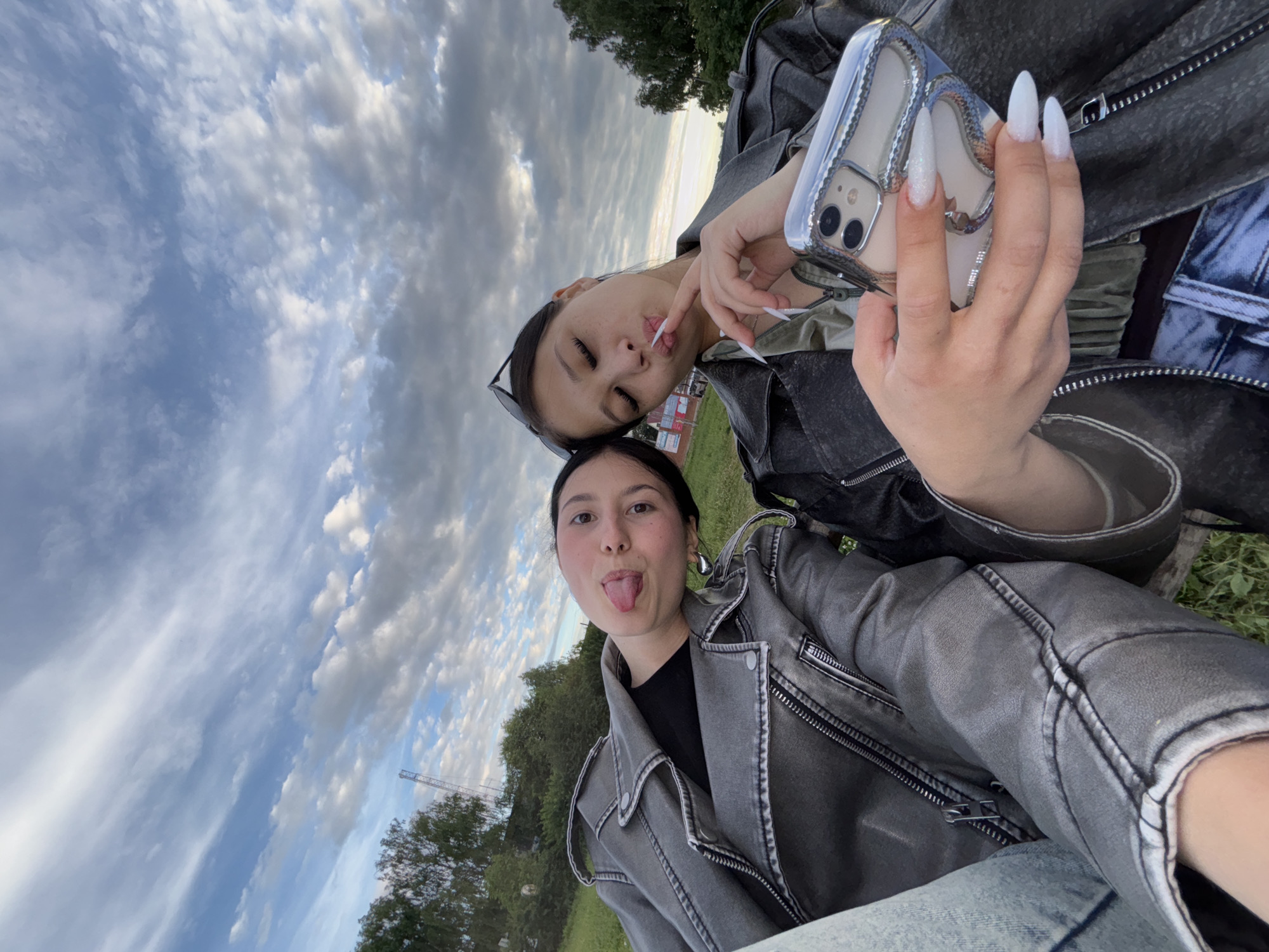 Two young women are posing for a selfie outdoors. The woman on the left has her tongue sticking out. The woman on the right is holding a phone and touching her lips with her finger. Both are wearing leather jackets. The background shows trees and a cloudy sky. The image appears to be taken in the city of Fokino, Russia, as indicated by context clues not visible in the image itself.