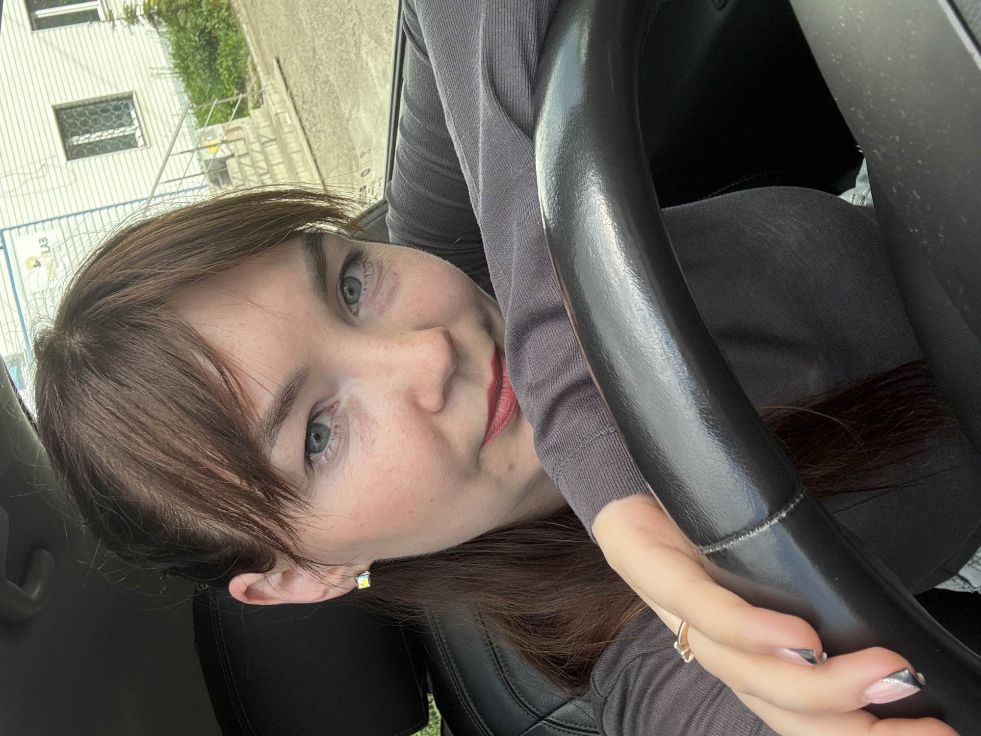A young woman with brown hair, blue eyes, and light skin is sitting in a car. She is wearing a grey long-sleeved shirt and a gold ring on her left hand. Her left arm is resting on the steering wheel, and her right arm is extended across the dashboard. Her fingernails are painted pink with silver tips. She has on small, square, iridescent earrings. The background shows a white building with windows and stairs, and some green foliage. The text on the building is partially obscured, but appears to read "CHAIKELAB." This suggests the location is likely Chaikovsky, Russia.
