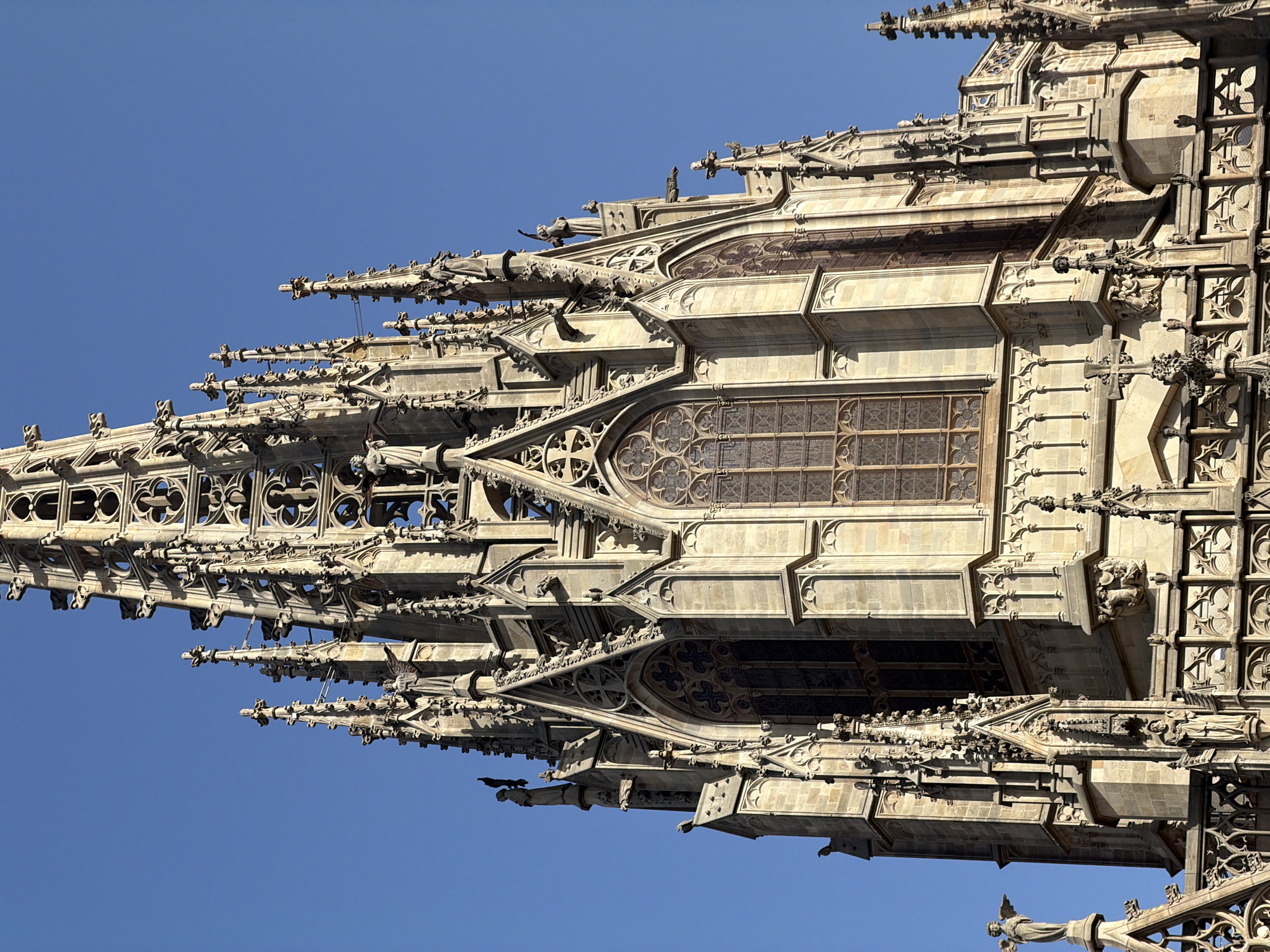 The image shows a close-up, low-angle shot of the intricate spires and facade of the Barcelona Cathedral (Catedral de Barcelona), a Gothic Roman Catholic cathedral in Barcelona, Spain. The sky is a clear, vibrant blue. The stone architecture is detailed with pointed arches, ornate carvings, gargoyles, and small statues of figures, including an angel on the left side of the image. Large, stained-glass windows with tracery are visible on the facade. The sunlight casts distinct shadows, highlighting the architectural details and creating a sense of depth. The composition focuses solely on the upper portions of the cathedral, emphasizing its verticality and elaborate design.