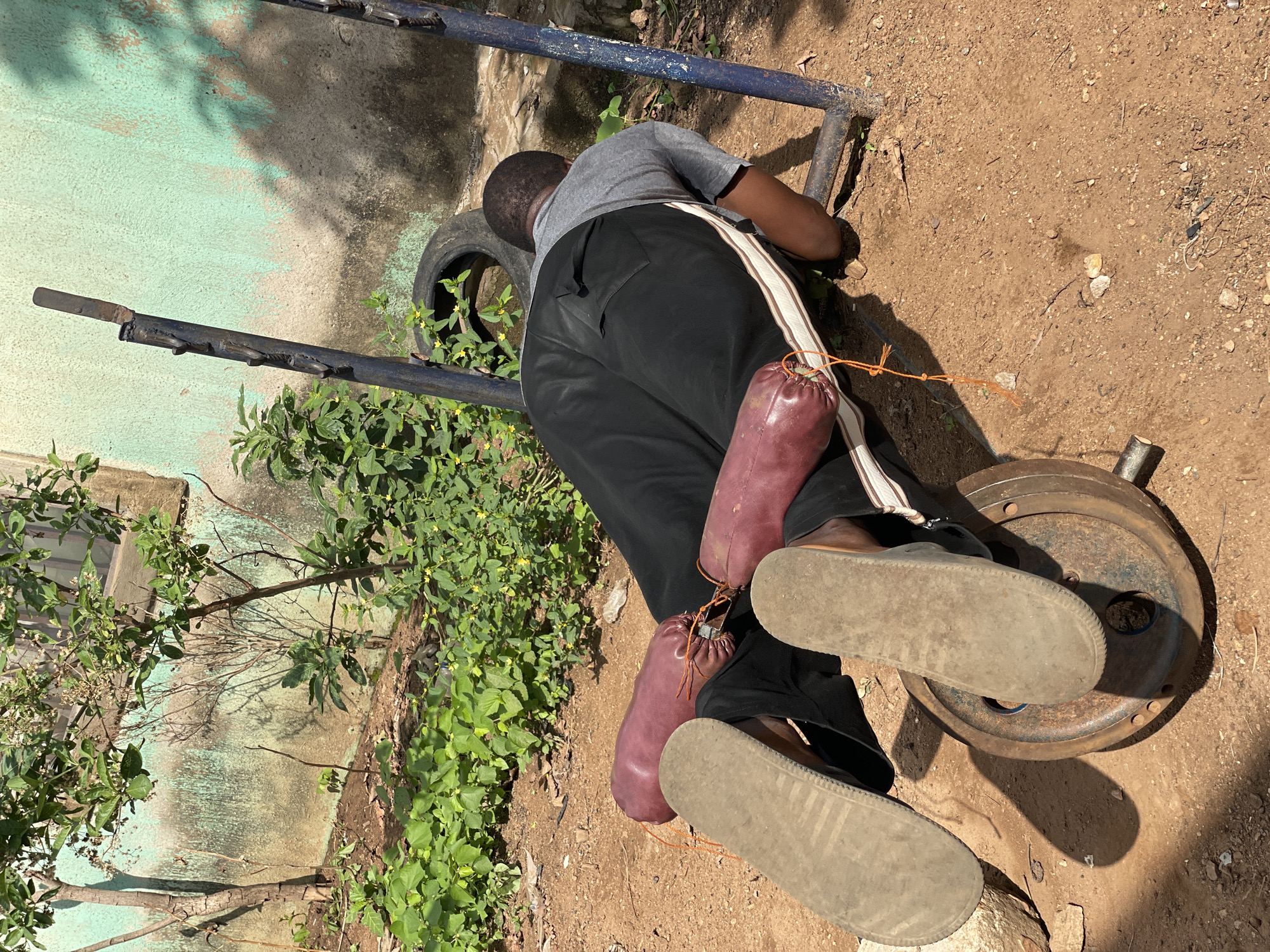 A person wearing a grey shirt, black pants with a white stripe, and sandals is lying face down on the ground in Akwanga, Nigeria. They appear to be working on a piece of exercise equipment constructed from metal tubing and a tire. A weight plate sits near the person's feet. The person is also carrying two burgundy-colored sacks with orange drawstrings. The ground is dirt and gravel, and there is a wall and some vegetation in the background.