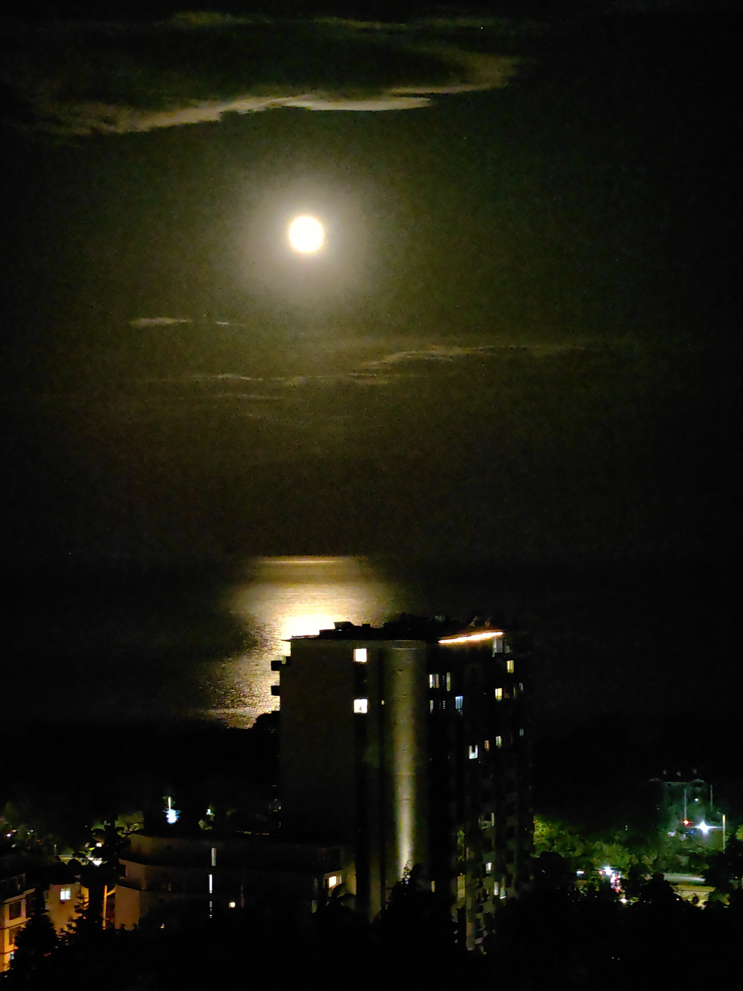 The image shows a night view in an unidentified city. A full moon is prominently featured in the sky, partially obscured by thin clouds, casting a bright reflection on a body of water below. The water's surface is illuminated along a path directly beneath the moon.
In the foreground, a multi-story building stands with several illuminated windows, suggesting occupancy. The building is flanked by trees and other structures, which are largely obscured by darkness. Streetlights and other illuminated signs are visible in the background, indicating urban development.